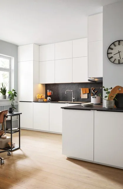 Modern white kitchen with black countertops, a clock on the wall, and plants near a window.