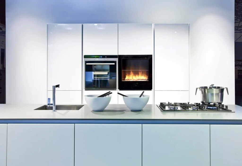 Modern kitchen with white cabinets, a built-in oven, a fireplace, a stovetop, a stainless steel pot, and two white bowls with utensils.