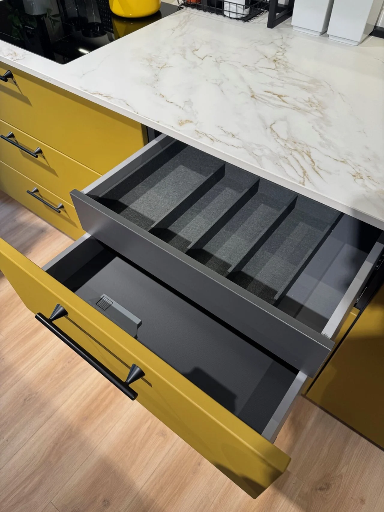 Open kitchen drawer with a yellow front, showing a black and gray divided utensil tray inside, with kitchen counter and yellow cabinets above.