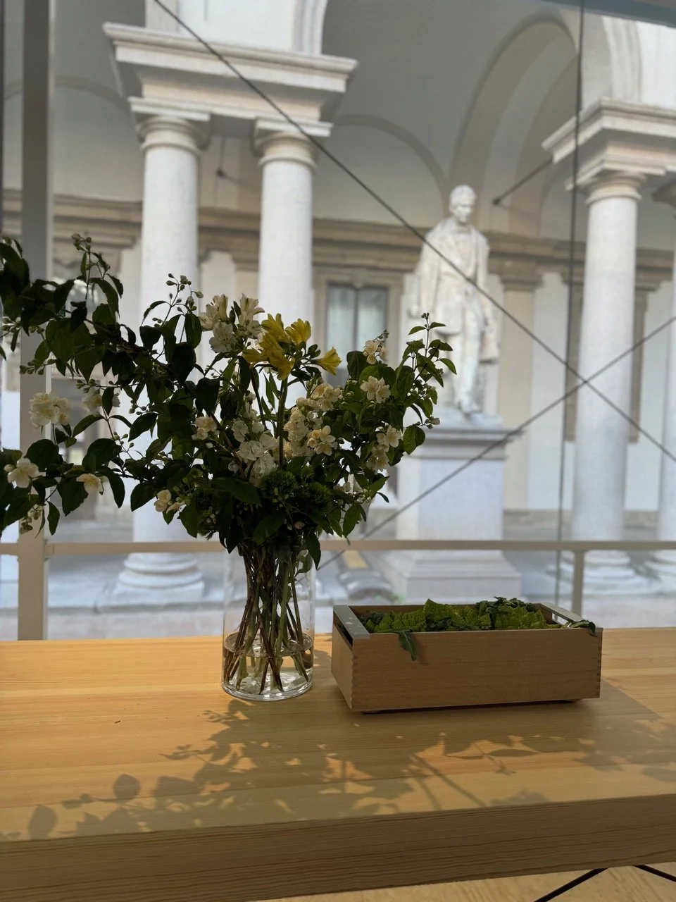 A glass vase with a bouquet of white and yellow flowers on a wooden table, with a wooden box containing green leaves beside it. A statue and classical columns are visible in the background through a window.