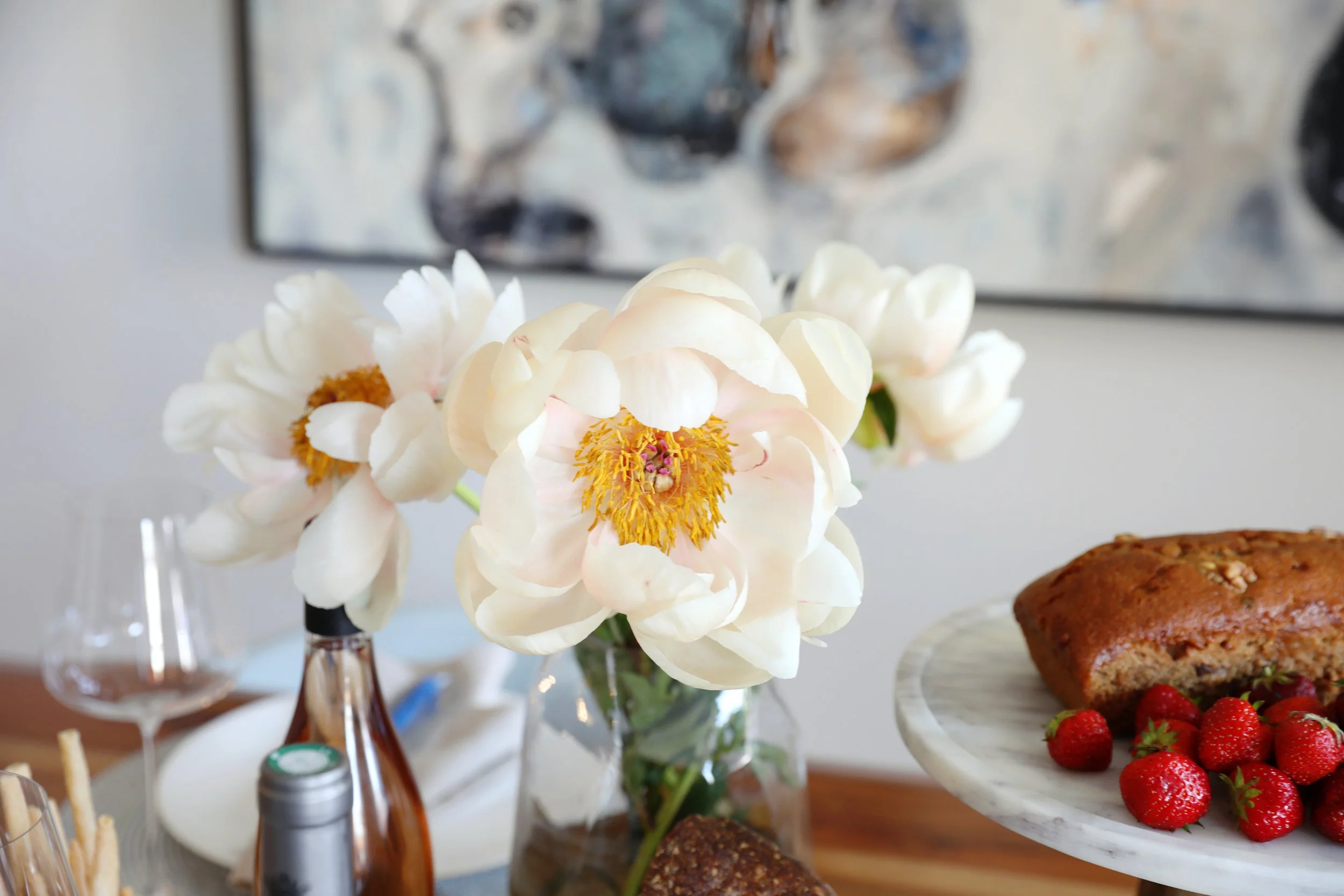 A close-up view of white peony flowers in a glass vase on a dining table, with a dessert and strawberries in the background.