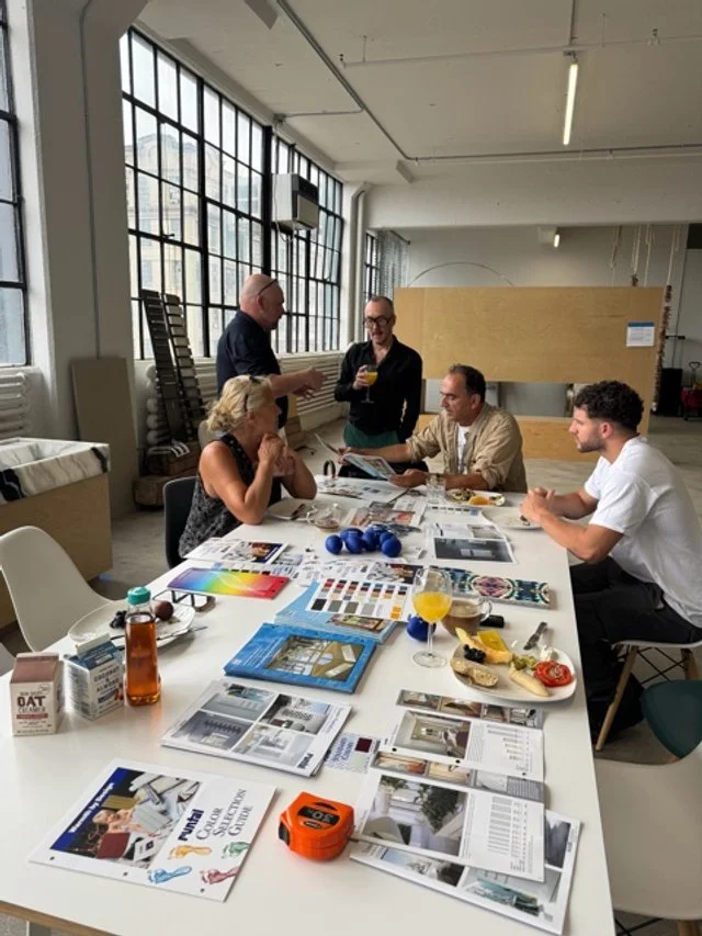 Five people engaged in a discussion around a table in a bright, modern office space with large windows, with design magazines, samples, and beverages on the table.