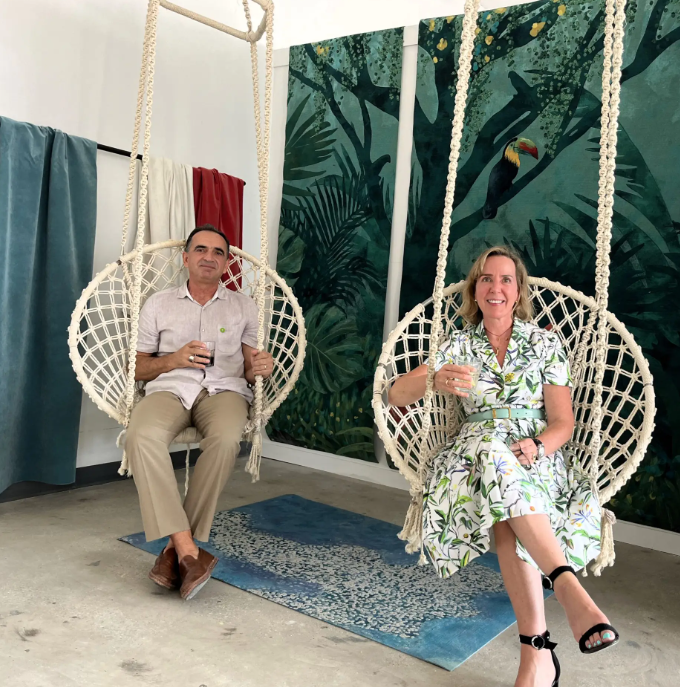 A man and woman sitting in hanging rope chairs, holding drinks, in front of a jungle-themed wall mural.