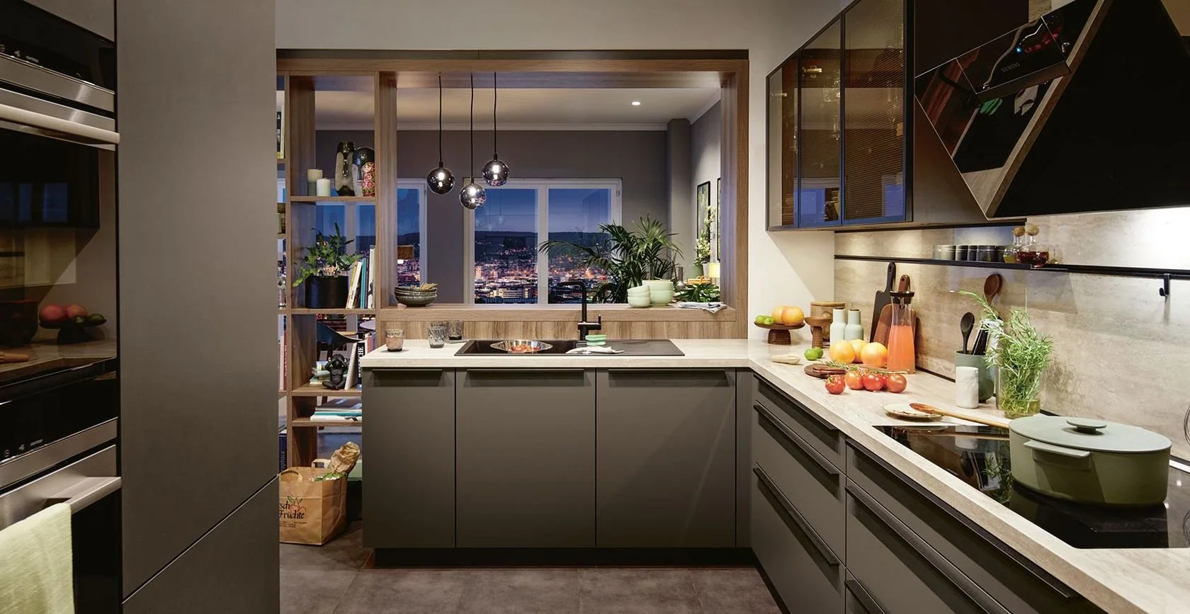 Modern kitchen with gray cabinets, beige countertops, cutting vegetables, fruit on counter, large window with city view, pendant lights, and open shelving.