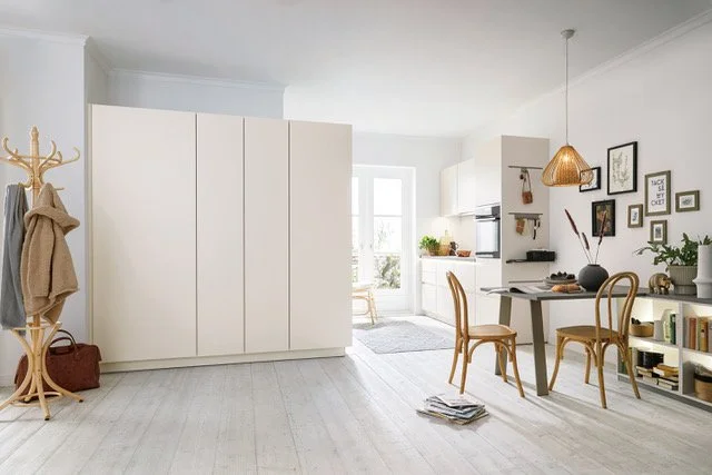 Bright, modern living space with white walls, a large white wardrobe, a cozy dining table with two chairs, and a gallery wall of framed photos and art. Natural light fills the room.