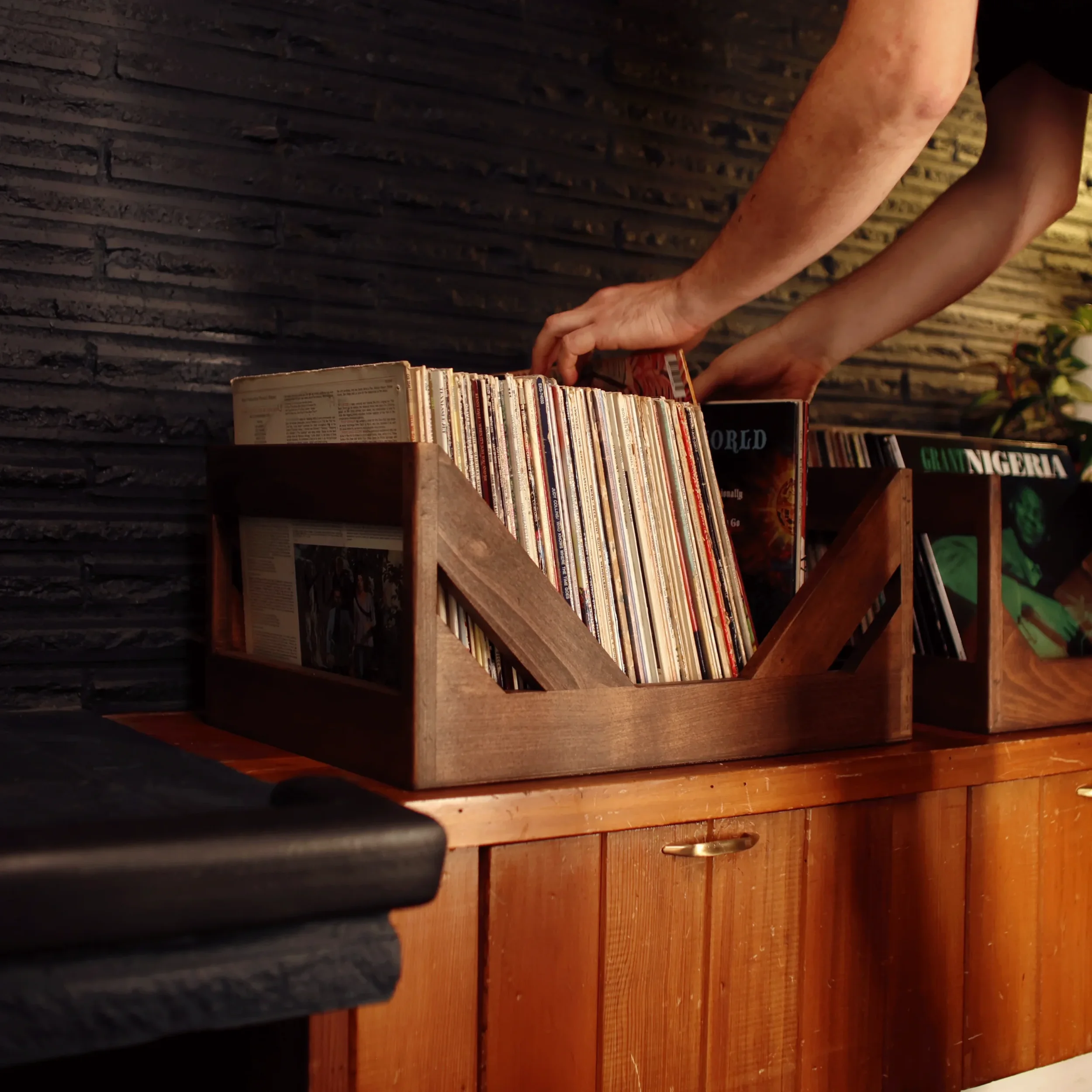 Person browsing vinyl records stored in a wooden crate on a wooden cabinet, with a textured dark wall in the background.