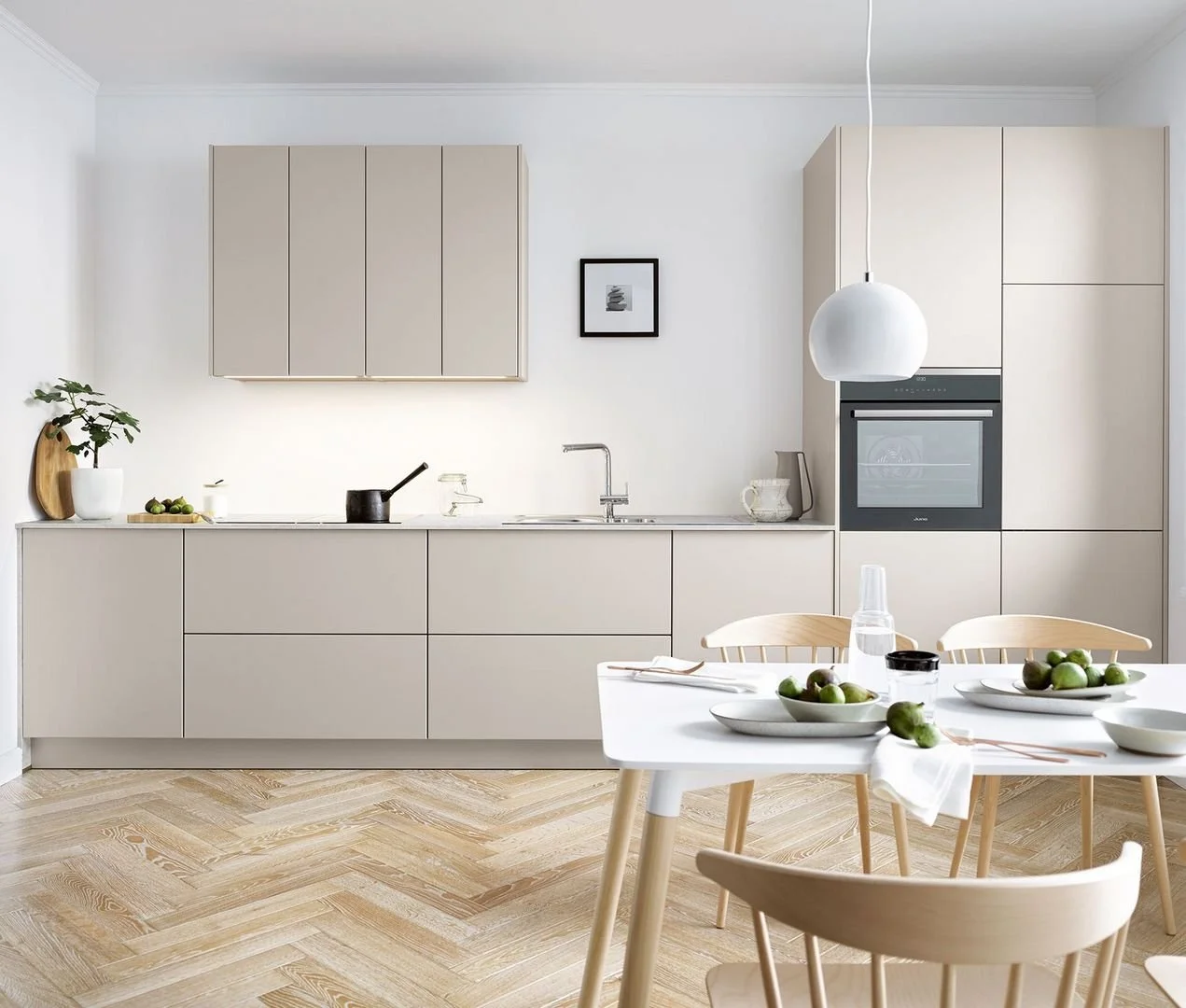 Minimalist kitchen with beige cabinets, light wood flooring, dining table with bowls of green apples, and a hanging white pendant light.