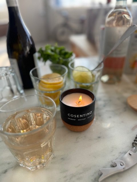 A candle labeled 'COSENTINO' burning on a table surrounded by glasses filled with drinks, slices of lemon or lime, a potted plant, a champagne or wine bottle, and various bar or kitchen items.