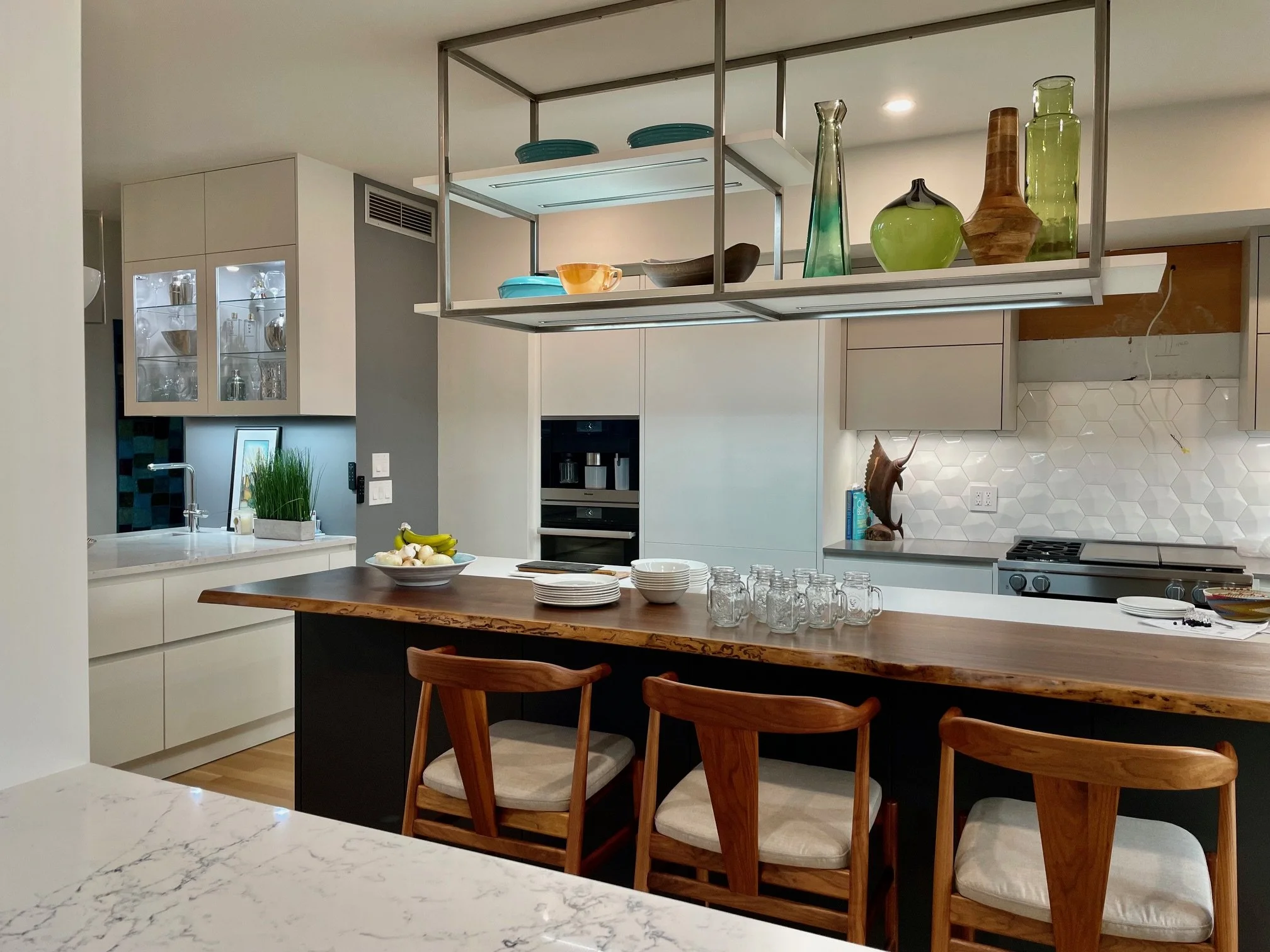 Modern kitchen with white cabinets, a marble countertop, a large wooden kitchen island with four wooden chairs, glass jars, and a decorative wooden fish sculpture. Open shelving with colorful vases and bowls, hexagonal tile backsplash, built-in oven, and a coffee machine.