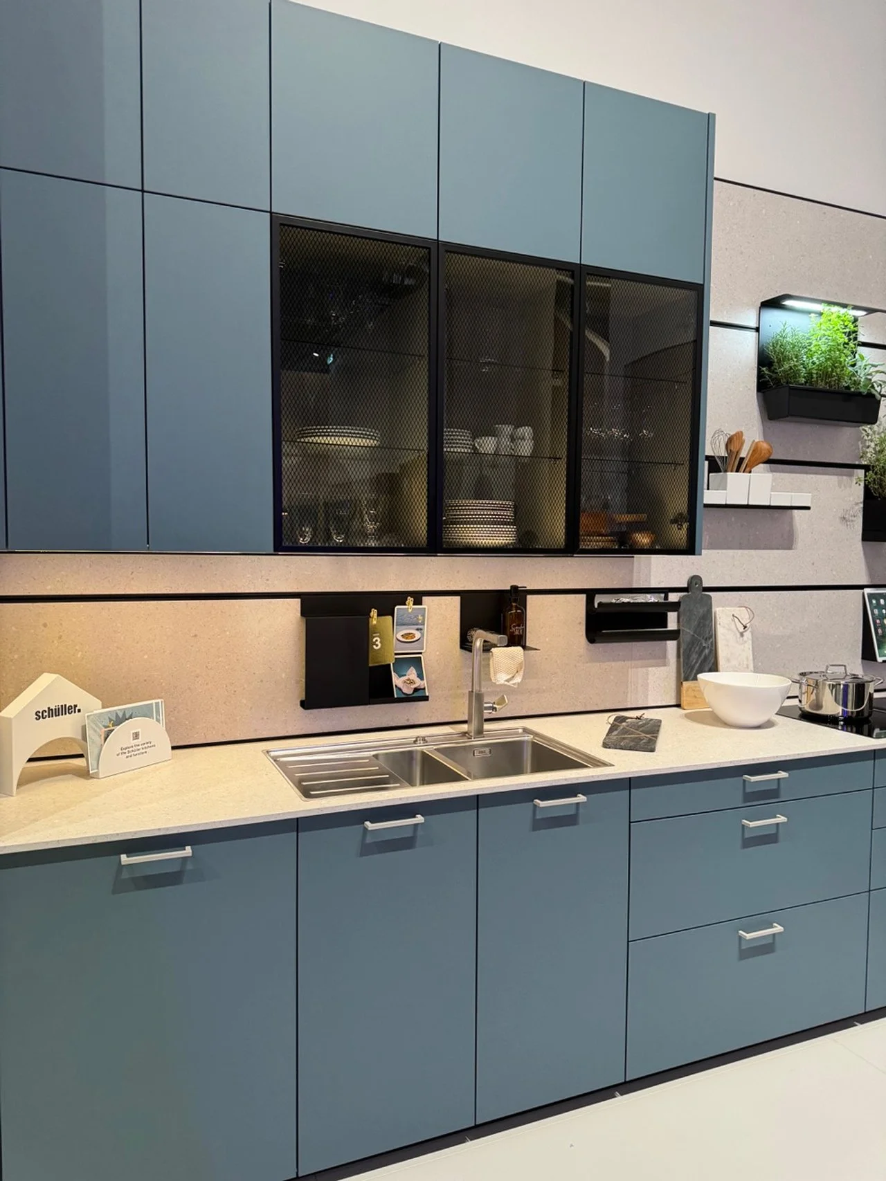 Modern kitchen with blue cabinets, white countertop, stainless steel sink, black and gray wall-mounted storage, and various kitchenware and plants.