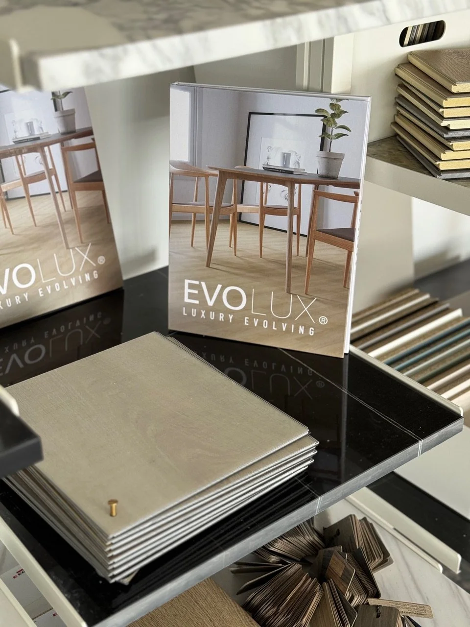 Interior display of flooring samples with a mirror showing a room scene featuring wooden chairs and a table, and a potted plant.
