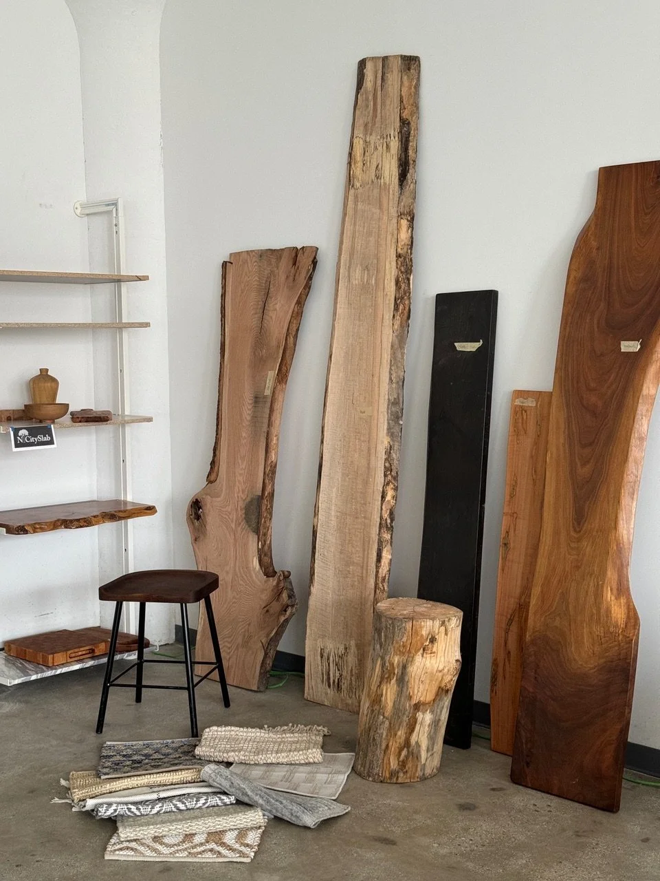 Display of wooden slabs and a log in a showroom with fabric samples on the floor and shelves with small items.