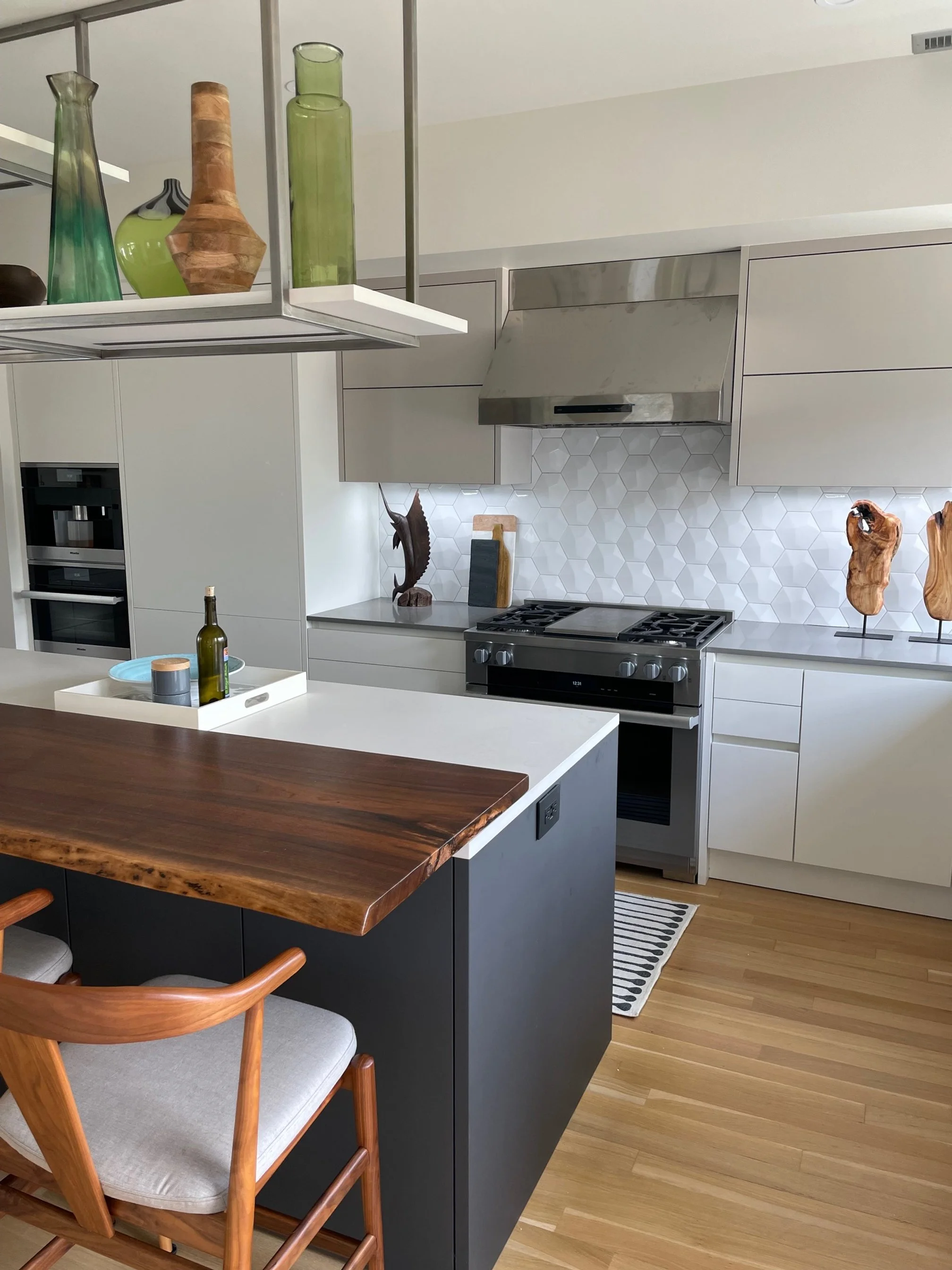 Modern kitchen with white cabinetry, stainless steel stove and range hood, decorative wood and ceramic art pieces on the counter, and a large kitchen island with a wood countertop and rustic wooden chairs, hardwood flooring, and a hanging fixture with decorative vases.