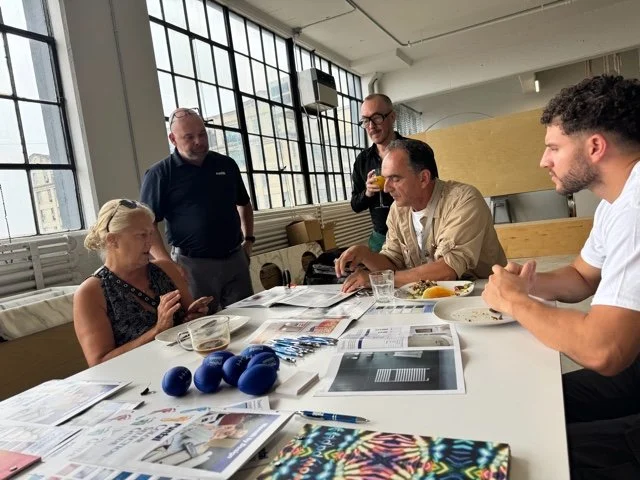 Group of five people in a meeting room with large windows, discussing and reviewing printed materials on a table, with some holding drinks.