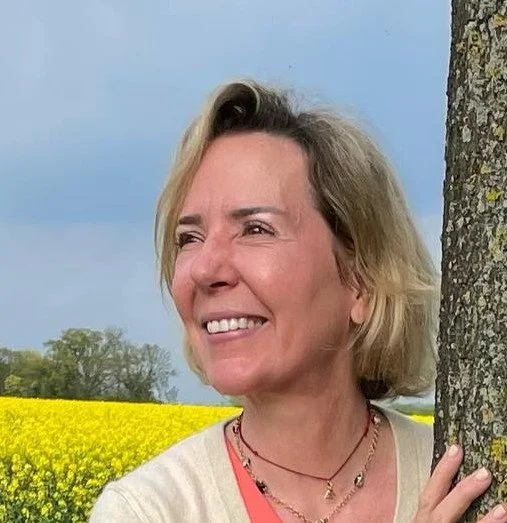 A woman with blonde hair smiling and leaning against a tree in a field of yellow flowers under a cloudy sky.