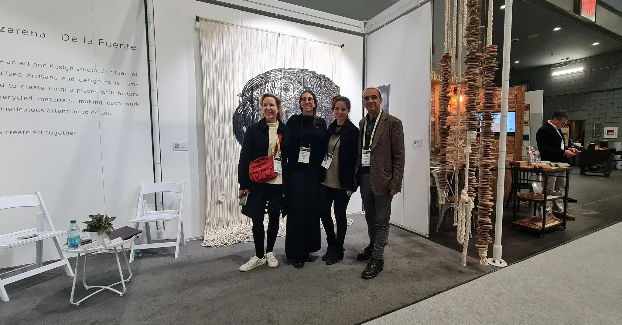 Four people standing together at an art display booth, smiling for the camera. The booth features a large wall hanging, a display of hanging circular objects resembling pottery or ceramics, and a sign mentioning art and design.