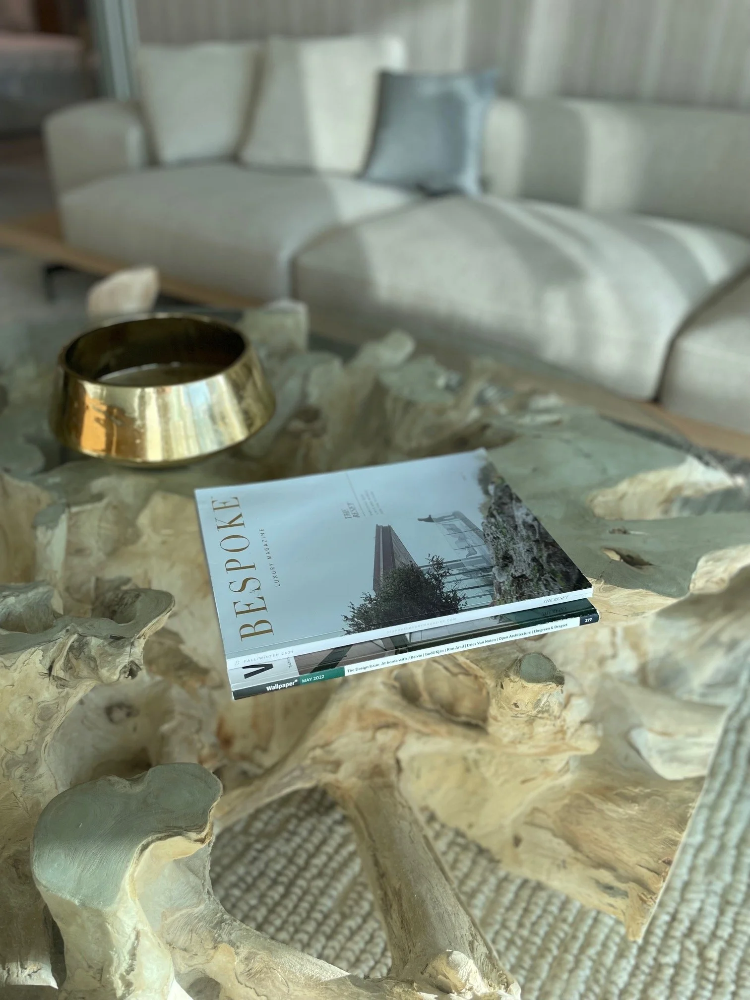 A magazine titled 'Bespoke' resting on a sculptural, natural wood table with a gold bowl nearby, in a cozy room with a white sofa in the background.