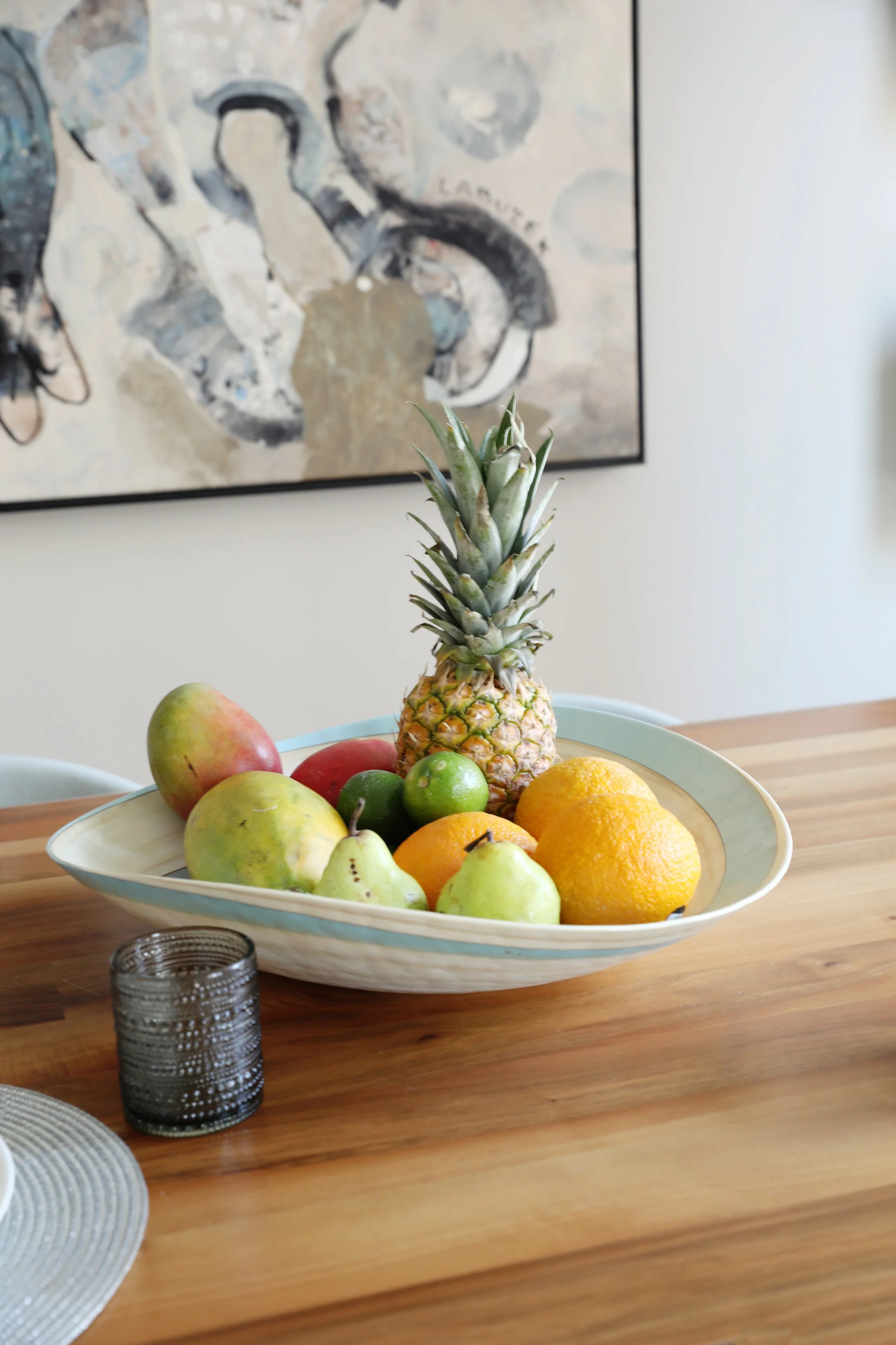 A white ceramic bowl filled with fresh fruits, including a pineapple, apples, pears, oranges, and limes, placed on a wooden table with a textured gray glass candle holder nearby, with abstract artwork on the wall in the background.