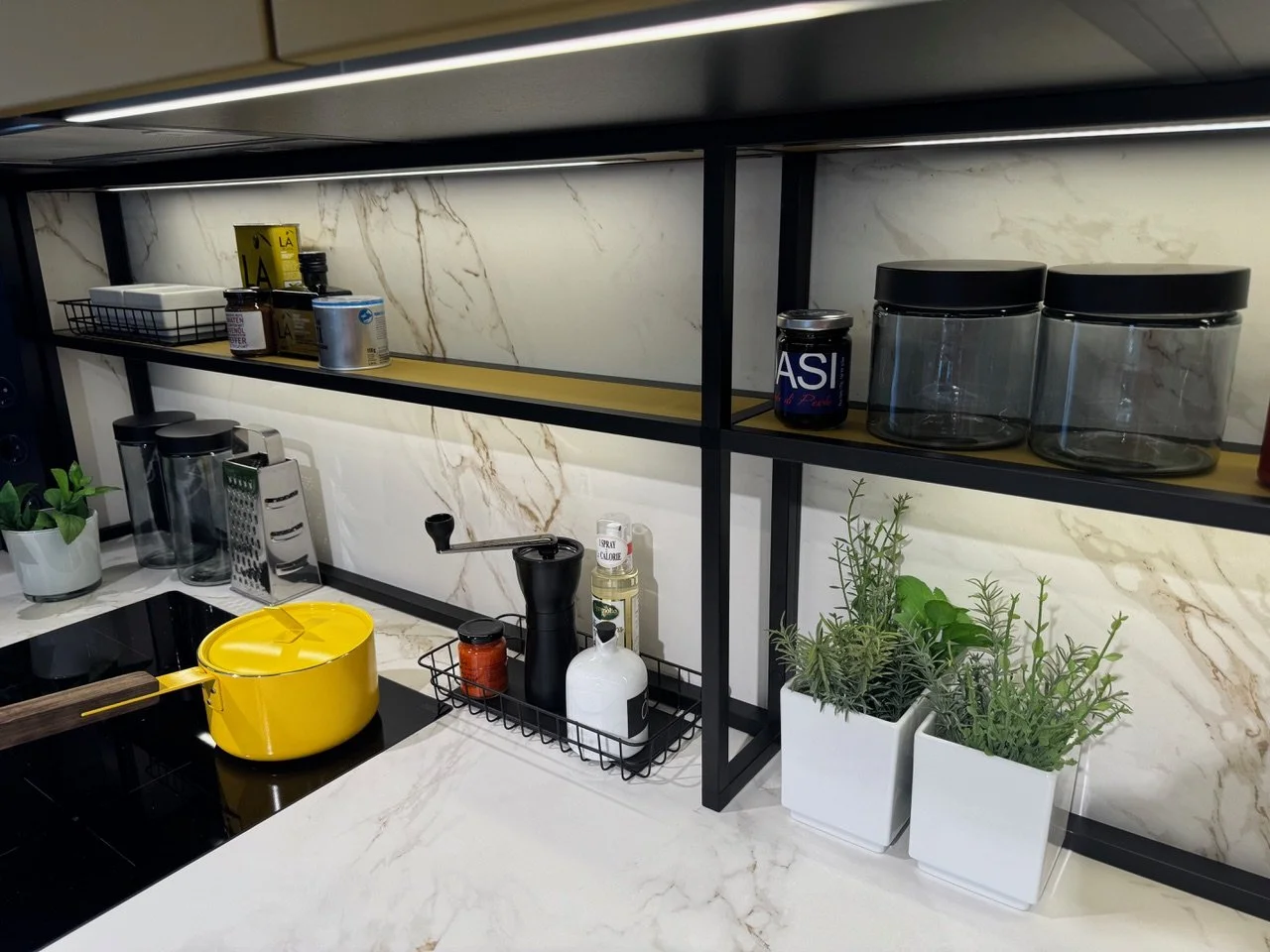 Modern kitchen countertop with various kitchen items, including a yellow pot, potted herbs, and glass jars on black shelving.