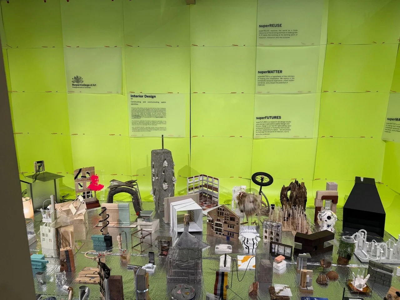A tabletop display of various miniature models of furniture, objects, and art pieces against a neon green background with informational posters.