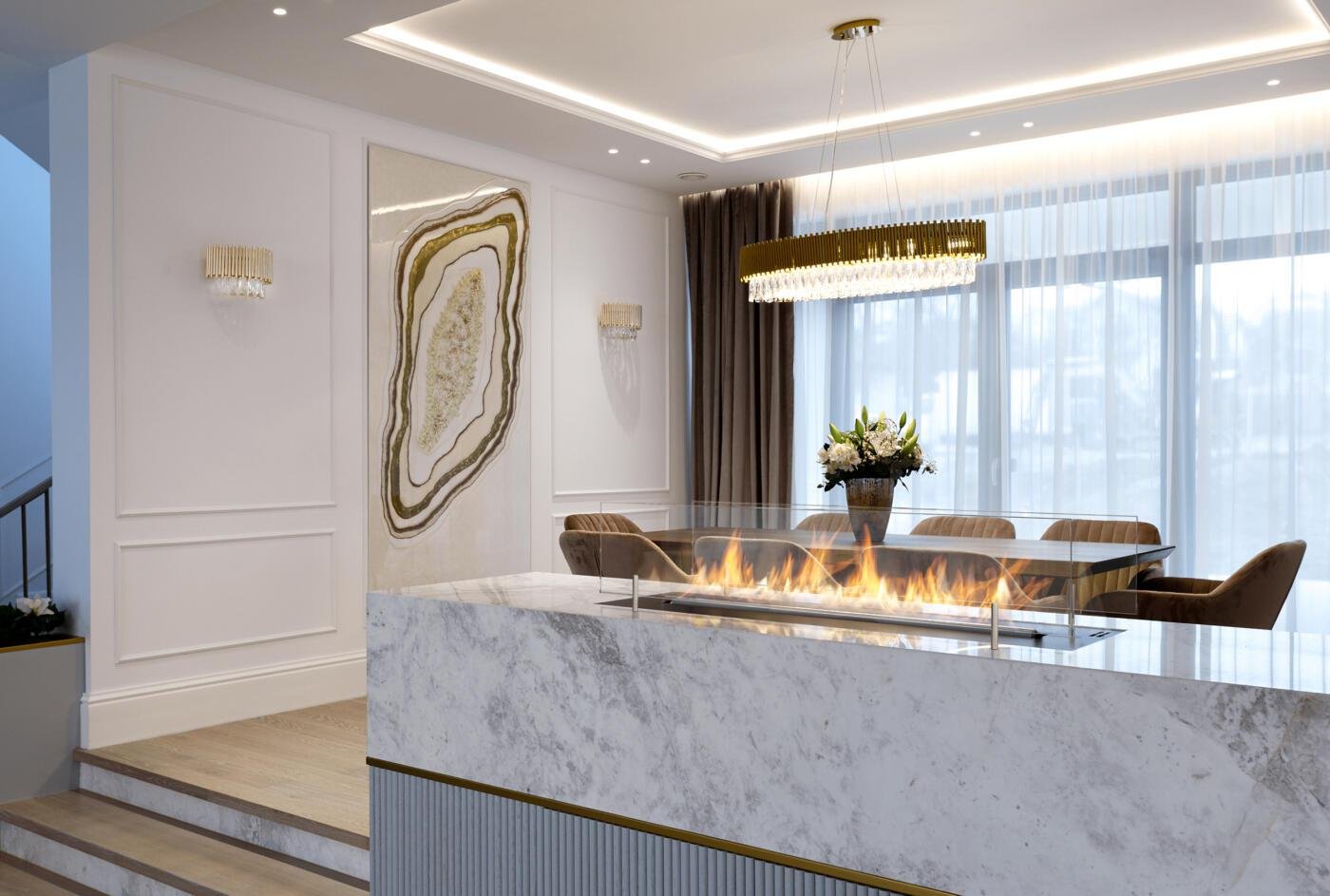 Modern dining room with a marble fireplace, a rectangular table surrounded by six beige upholstered chairs, a flower arrangement on the table, large window with sheer curtains, a gold chandelier, and wall sconces, with a staircase visible on the left.