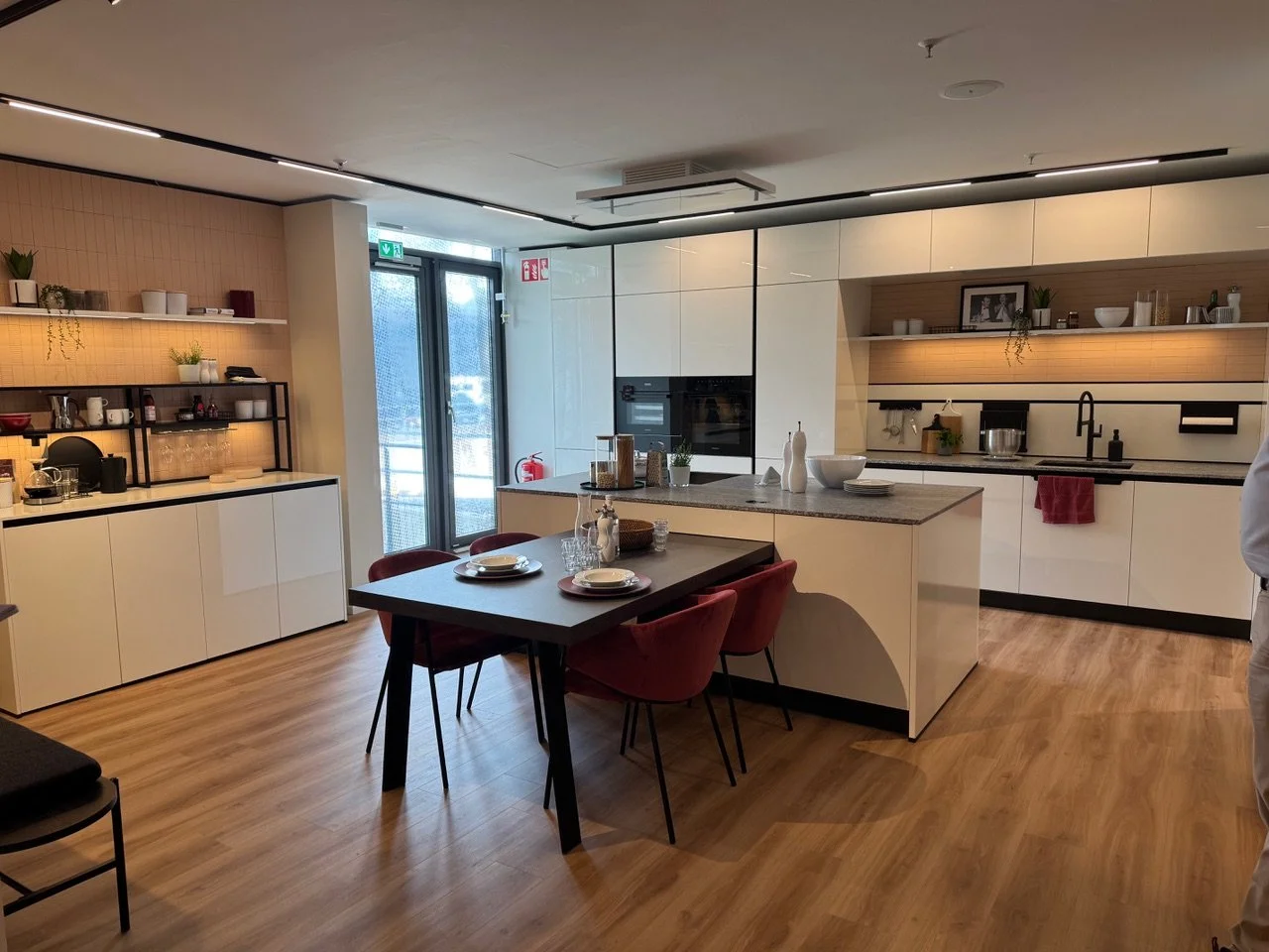 Modern kitchen with white cabinets, wooden flooring, a black dining table with red chairs, and open shelves with plants and kitchenware.