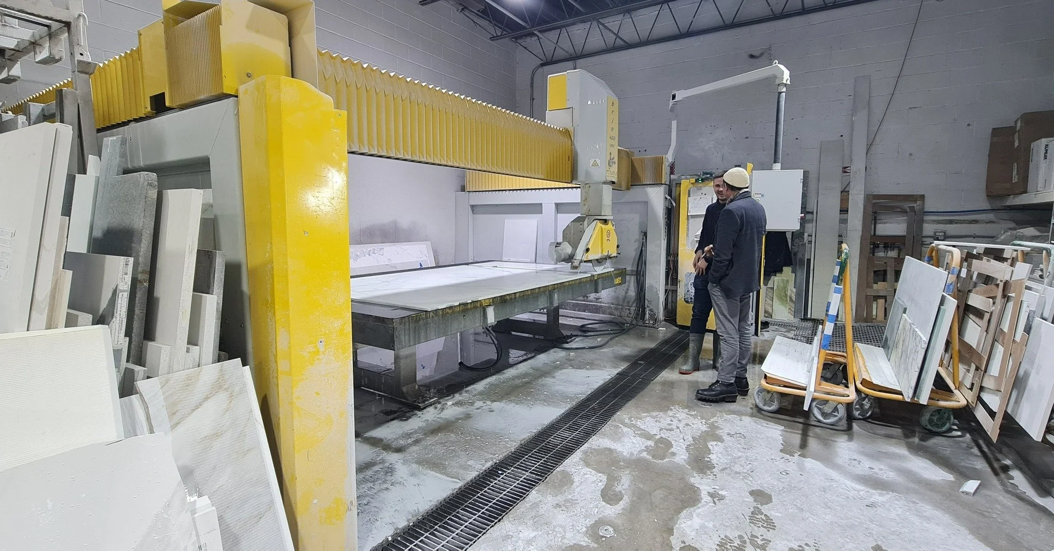Two men in the factory, one in a suit and the other in rubber boots, standing near a large industrial cutting machine with yellow accents, surrounded by slabs of stone or tile and carts holding similar materials.