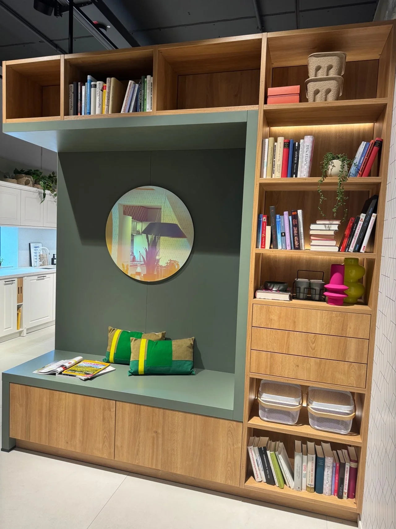 Wooden bookshelf with integrated sitting nook, containing books, decorative objects, and storage boxes, against a green wall with a round mirror.