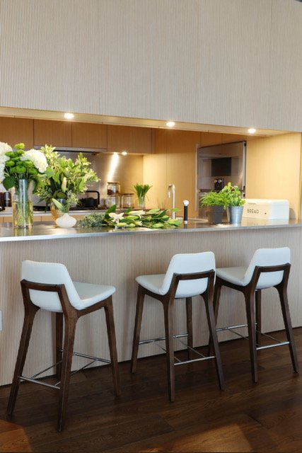 Modern kitchen with beige walls, a breakfast bar with three white and wood chairs, green plants and flowers on the counter, and kitchen appliances in the background.