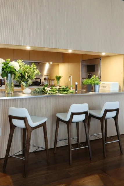 Modern kitchen with a breakfast bar, white chairs with wooden legs, fresh green plants, a white bread box, and a view into the kitchen area with cabinets and appliances.