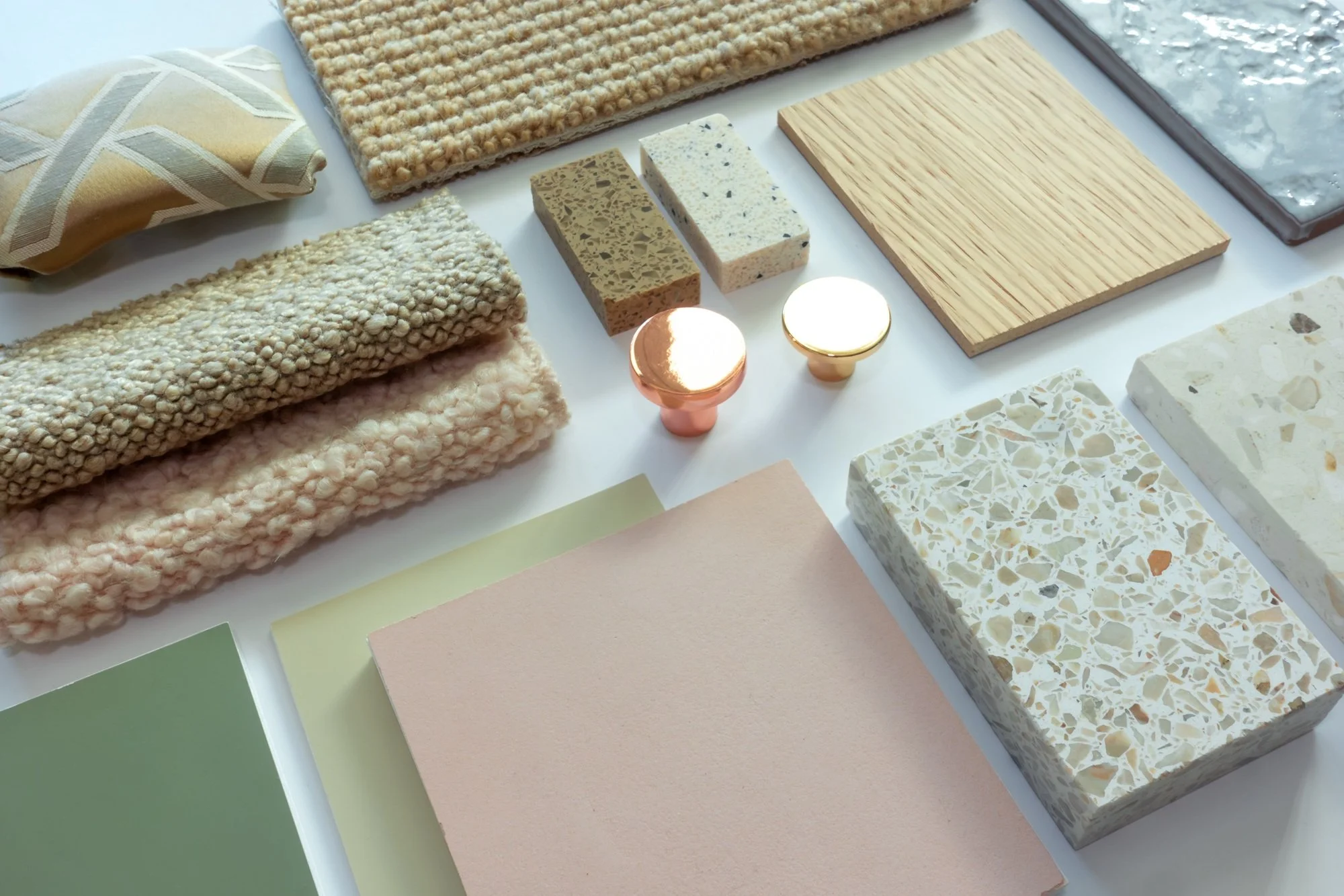 Display of various fabric swatches, tiles, and color samples on a table, including textured textiles, stone tiles, wood samples, and paper in pastel and neutral tones.