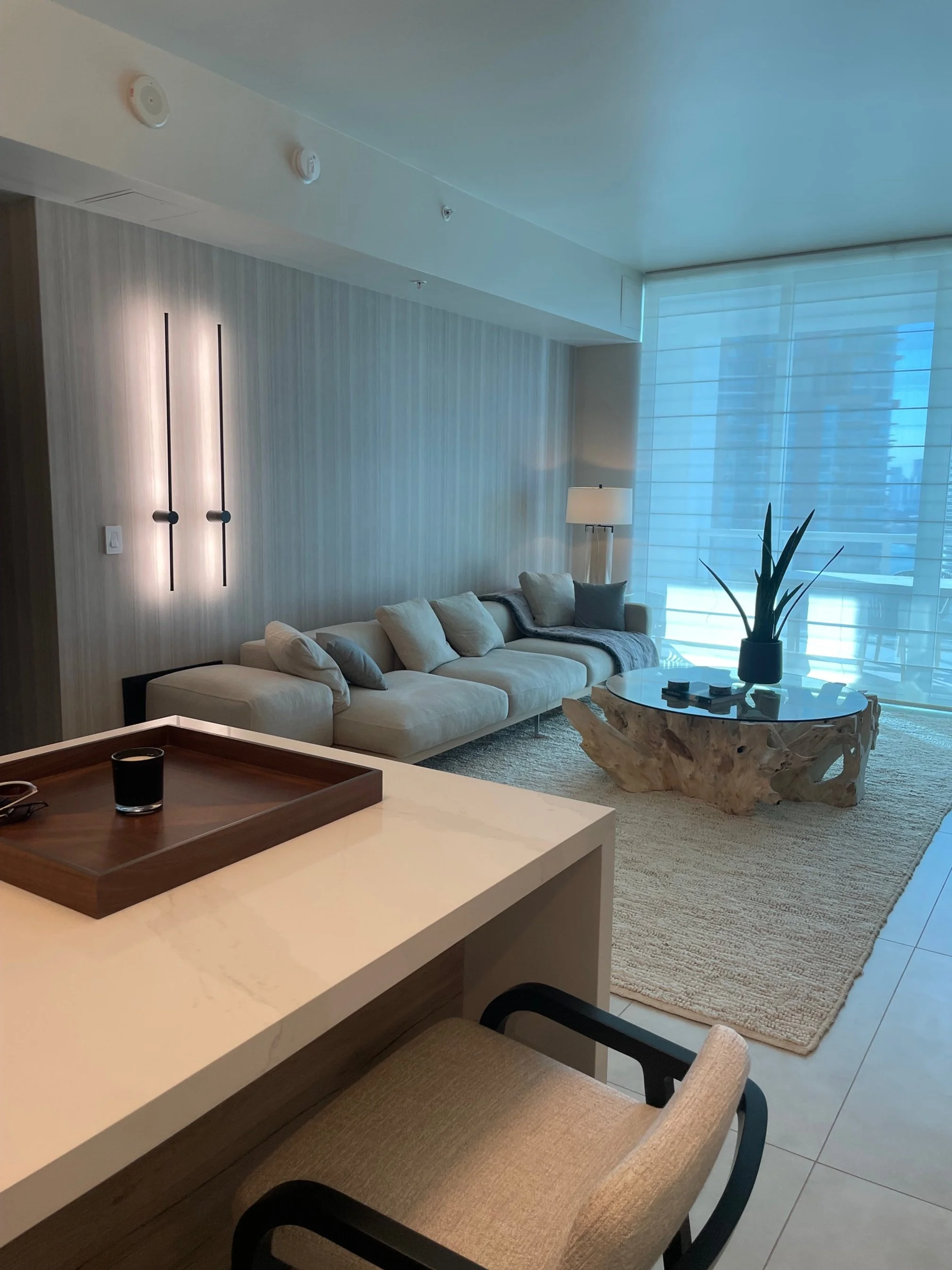 Living room with cream-colored sofa, wooden and marble coffee table, large window with sheer curtains, houseplant, and modern decor.