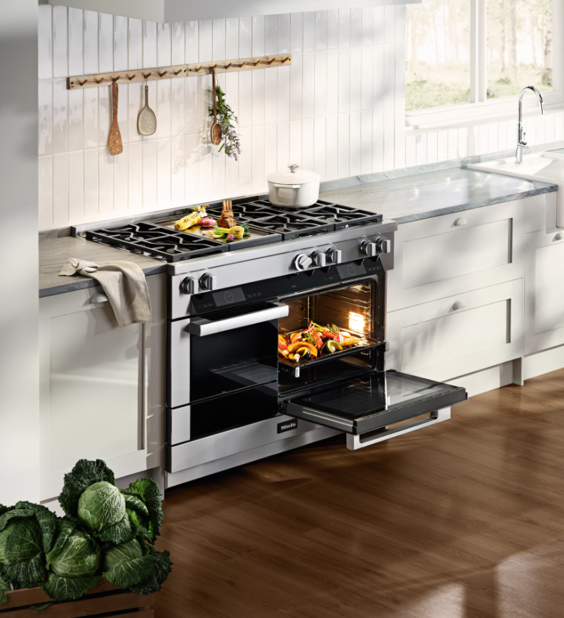 Kitchen scene with a modern stove oven, vegetables inside the oven, and fresh vegetables on the countertop, with a window and white cabinetry.