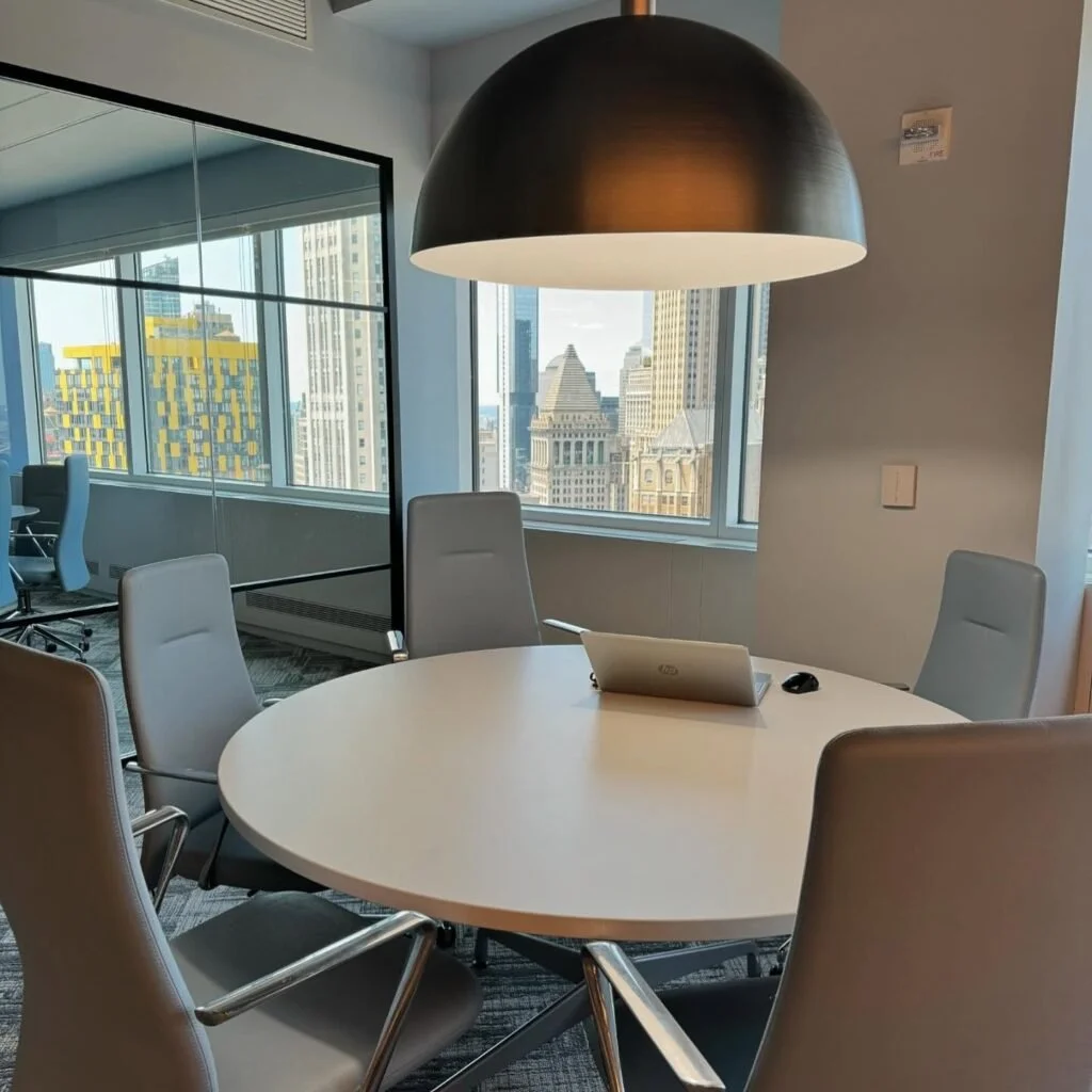 Conference room with a round table, five gray chairs, a black pendant lamp, a laptop, a mouse, large windows with a cityscape view including tall buildings.