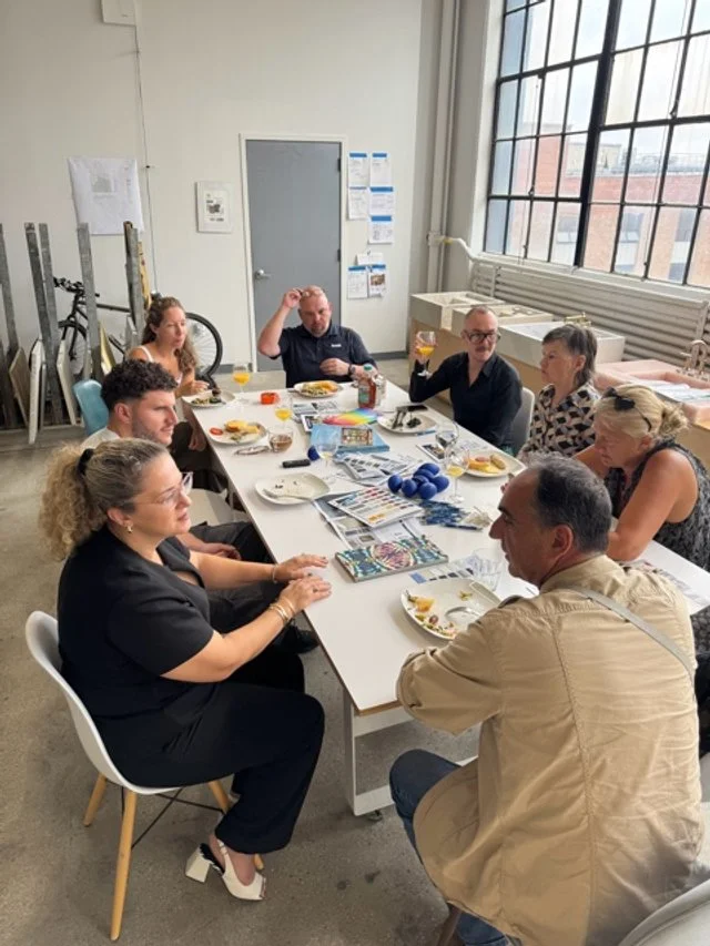 Group of seven people seated around a table during a meeting or dinner in a bright room with large windows and minimal decor.