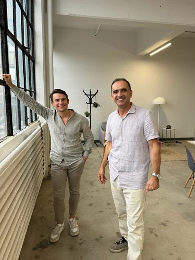 Two men standing in a modern office space with large windows, smiling at the camera.