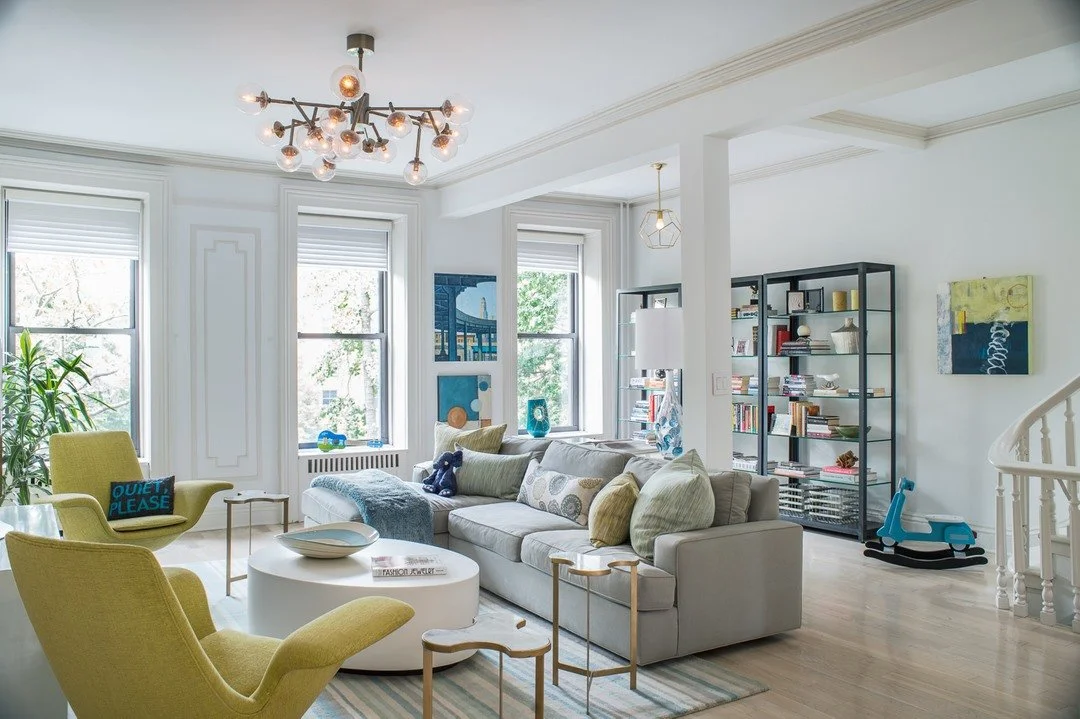 Bright living room with white walls, large windows, a beige sofa with colorful pillows, two yellow armchairs, a white coffee table, a modern chandelier, bookshelves with books and decor, and a blue rocking horse.