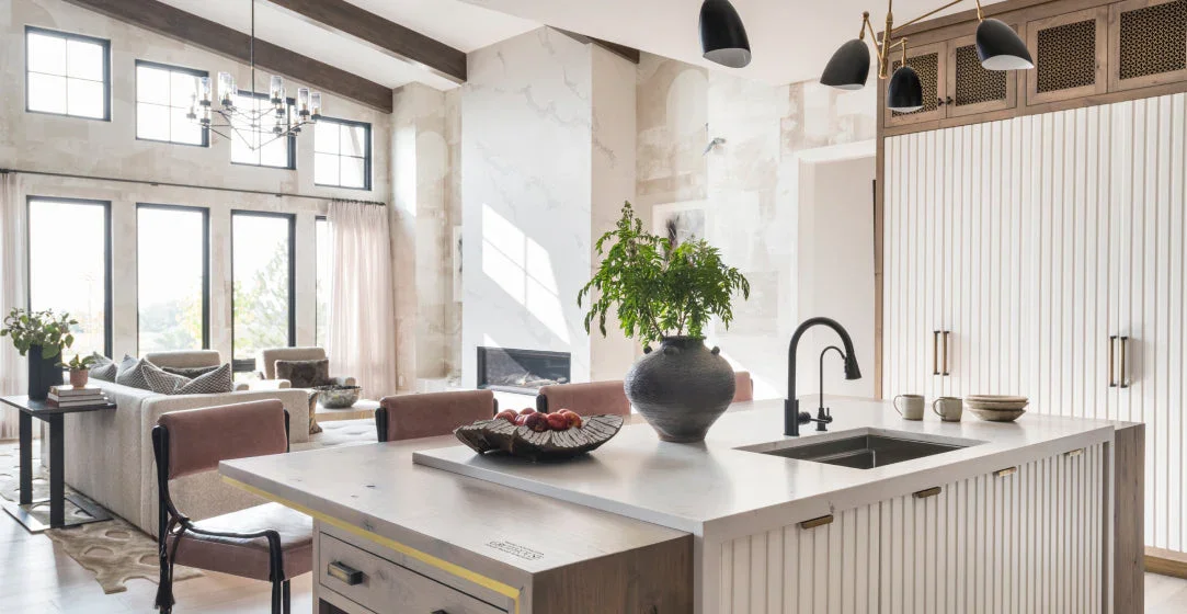 Open-concept living room and kitchen with large windows, modern chandelier, potted plants, and a kitchen island with a black faucet and a potted fern.