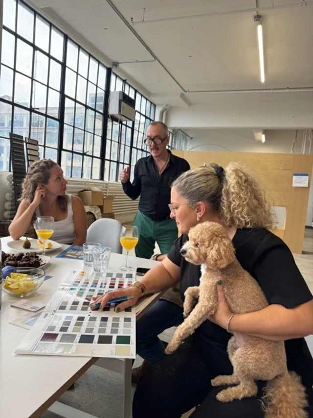 A group of three people and a dog sitting around a table in a well-lit room with large windows. The woman on the right holding a beige poodle, pointing at a color sample book. The man standing in the middle appears to be explaining something, while t