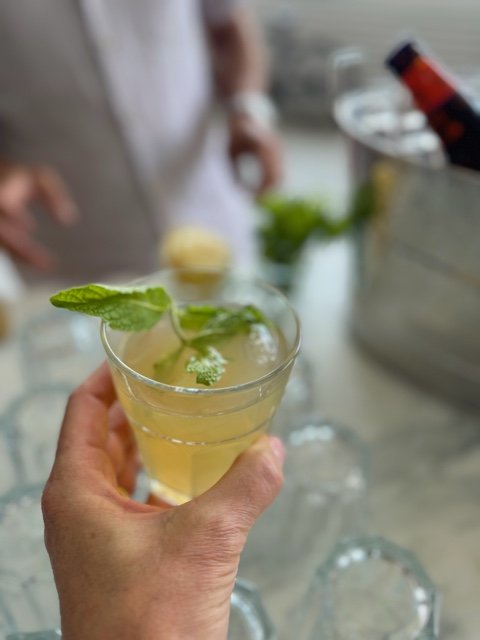 A person holding a glass of pale yellow cocktail garnished with a mint leaf.