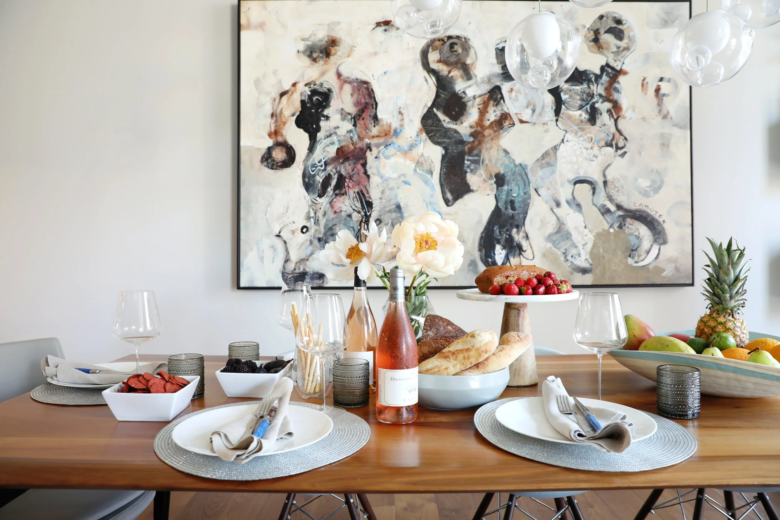 A table set for a meal with wine bottles, flowers, bread, fruits, and a cake, in front of an abstract black and white painting.