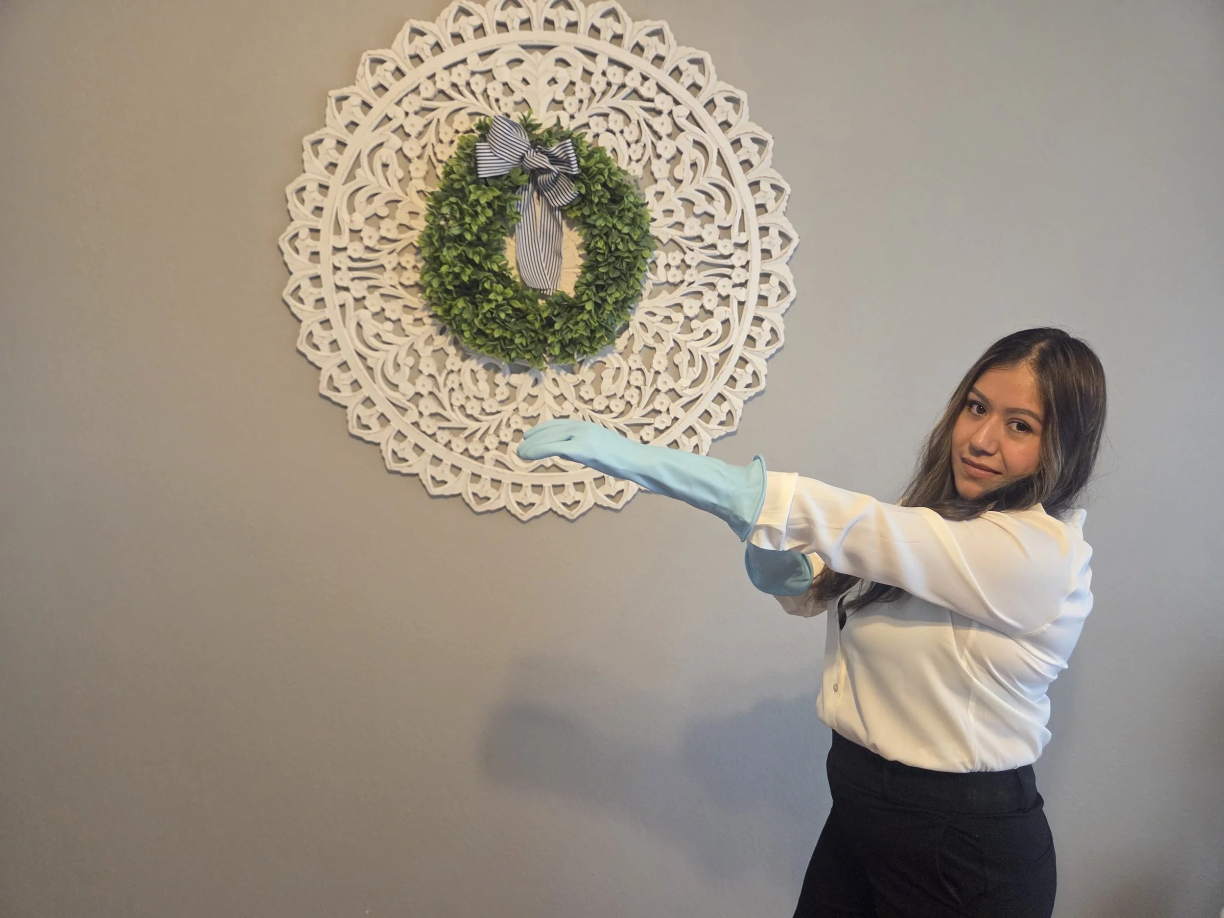 A woman with long dark hair wearing a white blouse and black pants is standing in front of a gray wall. She is pointing at a decorative wall piece—a white, intricate, circular wall hanging with a green wreath and a black-and-white striped bow on it.