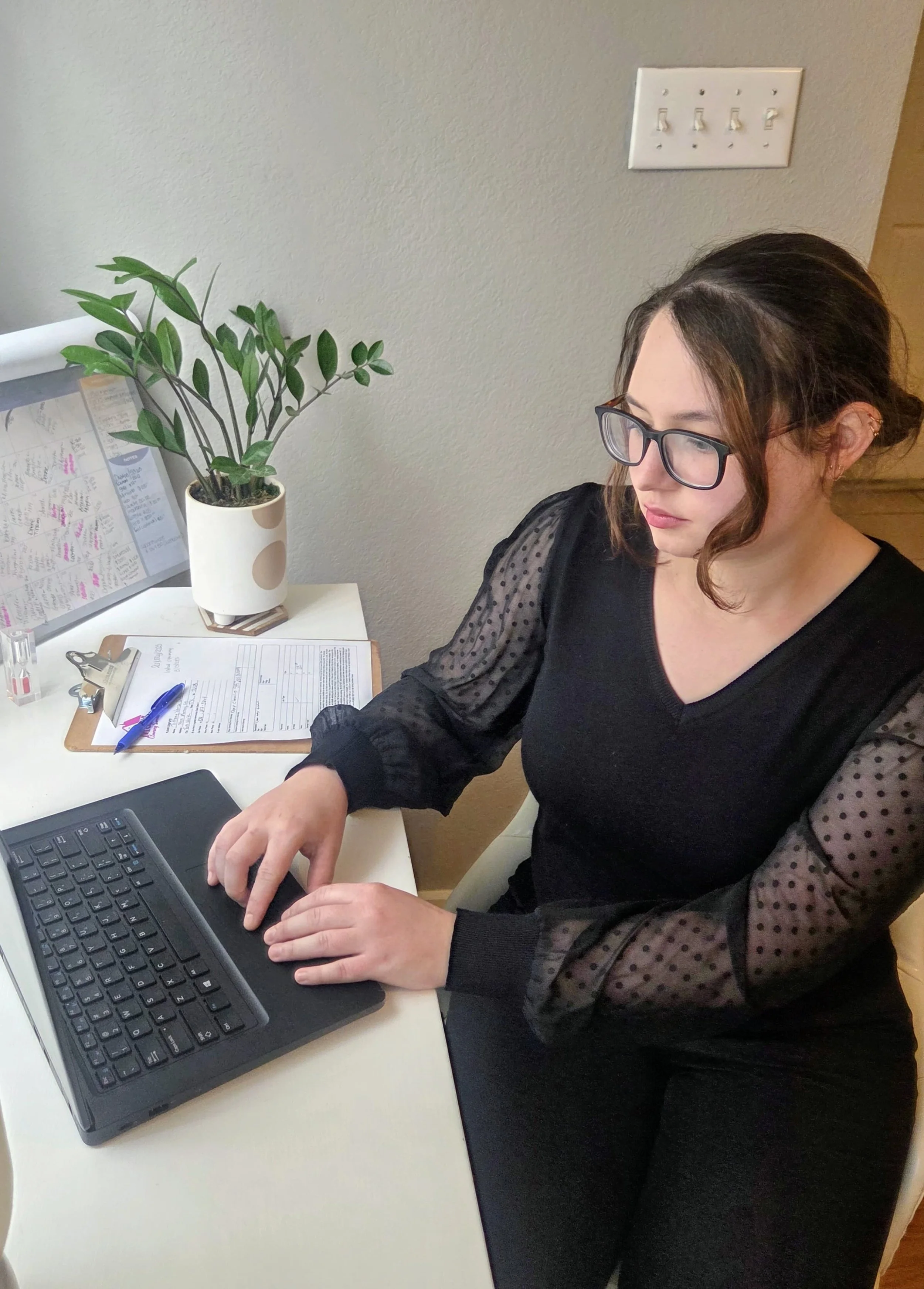 Woman in black blouse with sheer dotted sleeves working on a laptop at a white desk, with a potted plant, clipboard, and papers nearby.