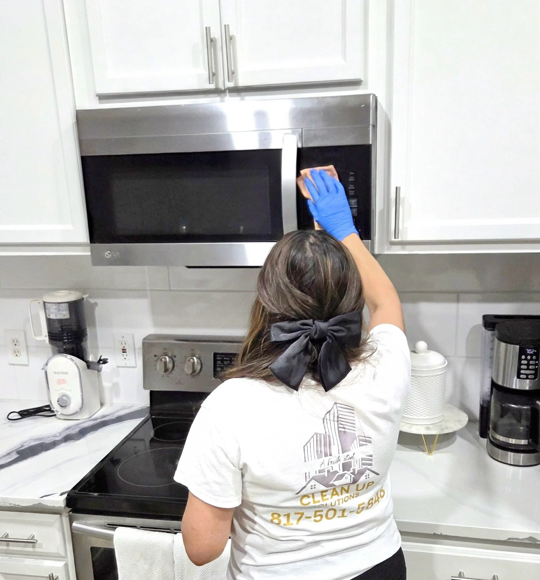 A woman cleaning a microwave oven in a kitchen, wearing a white t-shirt with a company logo and a black bow in her hair.