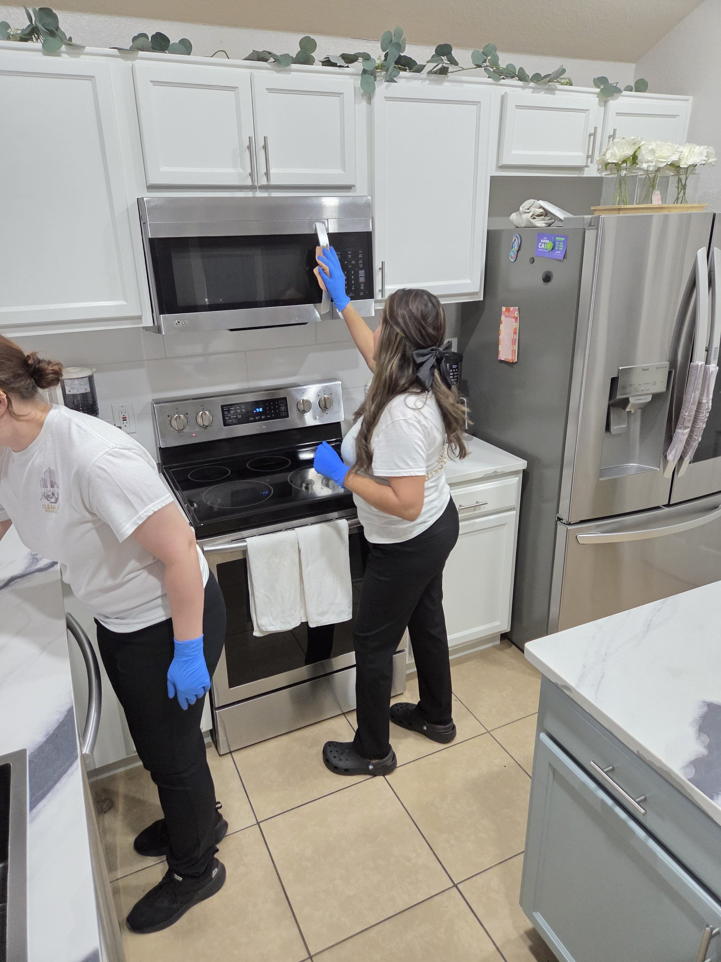 Two women cleaning a kitchen. One woman is reaching to clean the microwave, while the other is standing nearby cleaning a stove. Both are wearing blue gloves and casual clothing.