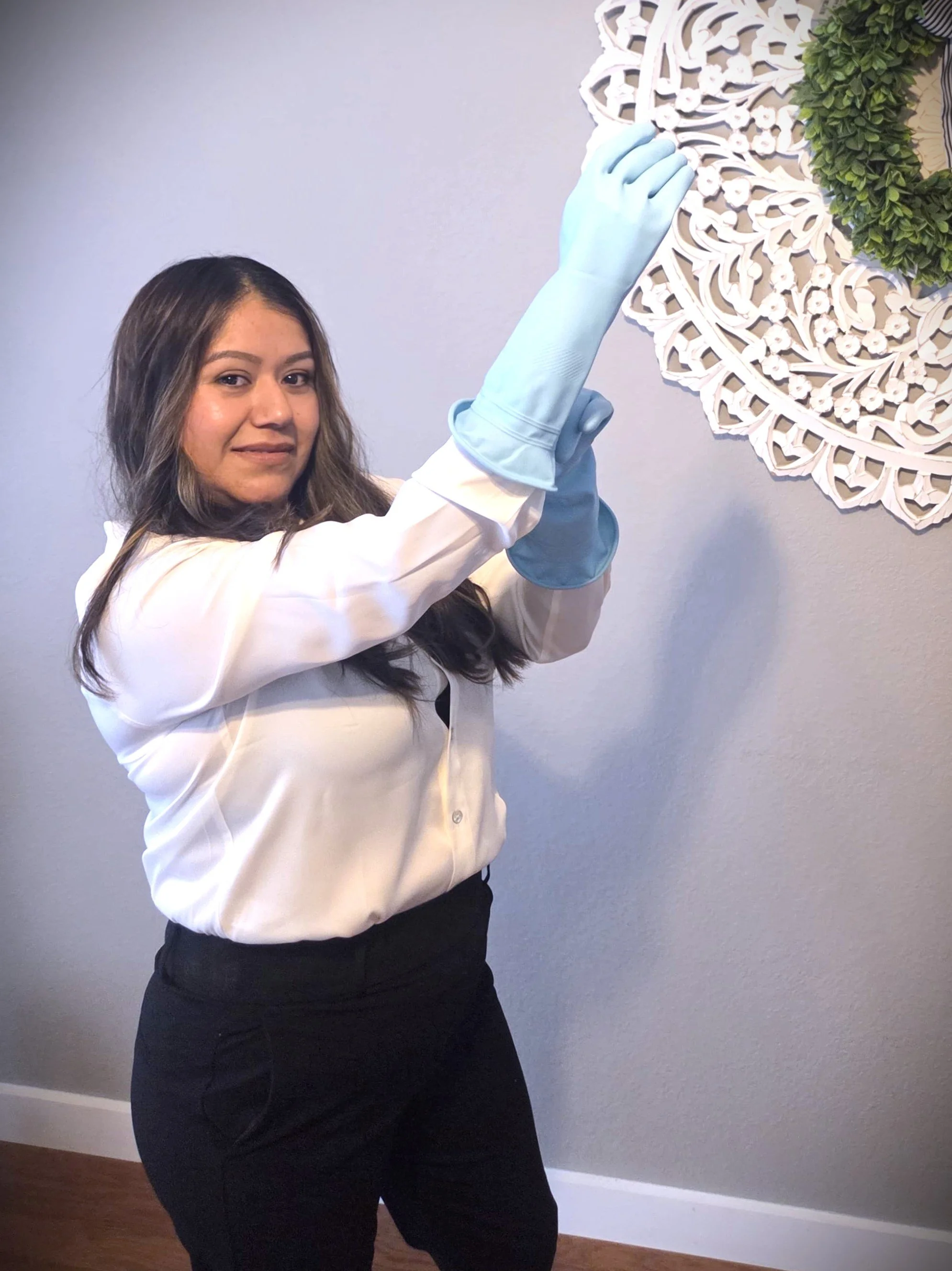 A woman wearing a white blouse and black pants is hanging a decorative white wooden wall art piece with a green wreath on a wall
