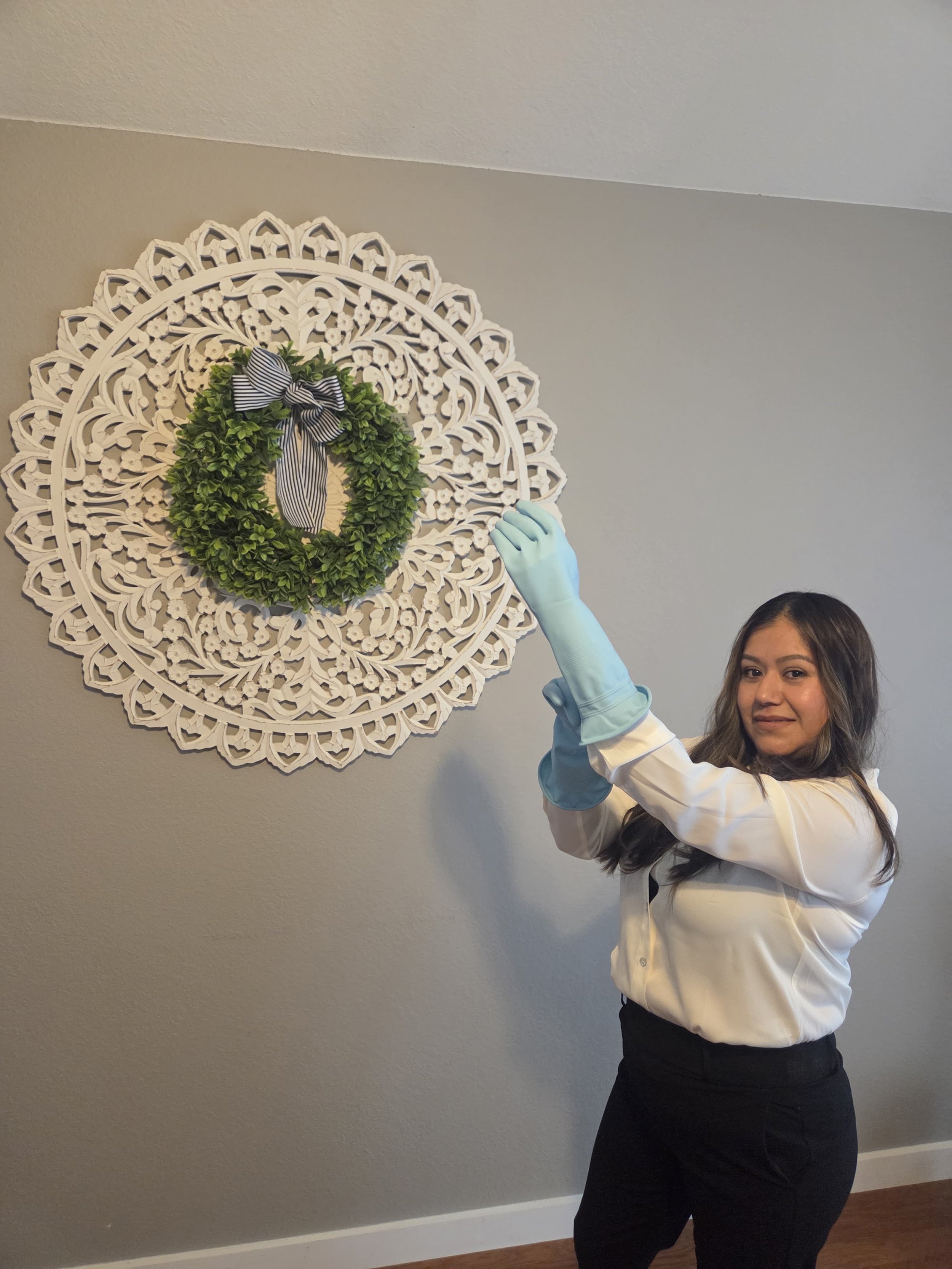 Woman wearing white shirt and black pants, standing near a wall, holding a pair of blue gloves, facing the camera, with a decorative white wreath with a black-and-white striped bow on the wall.