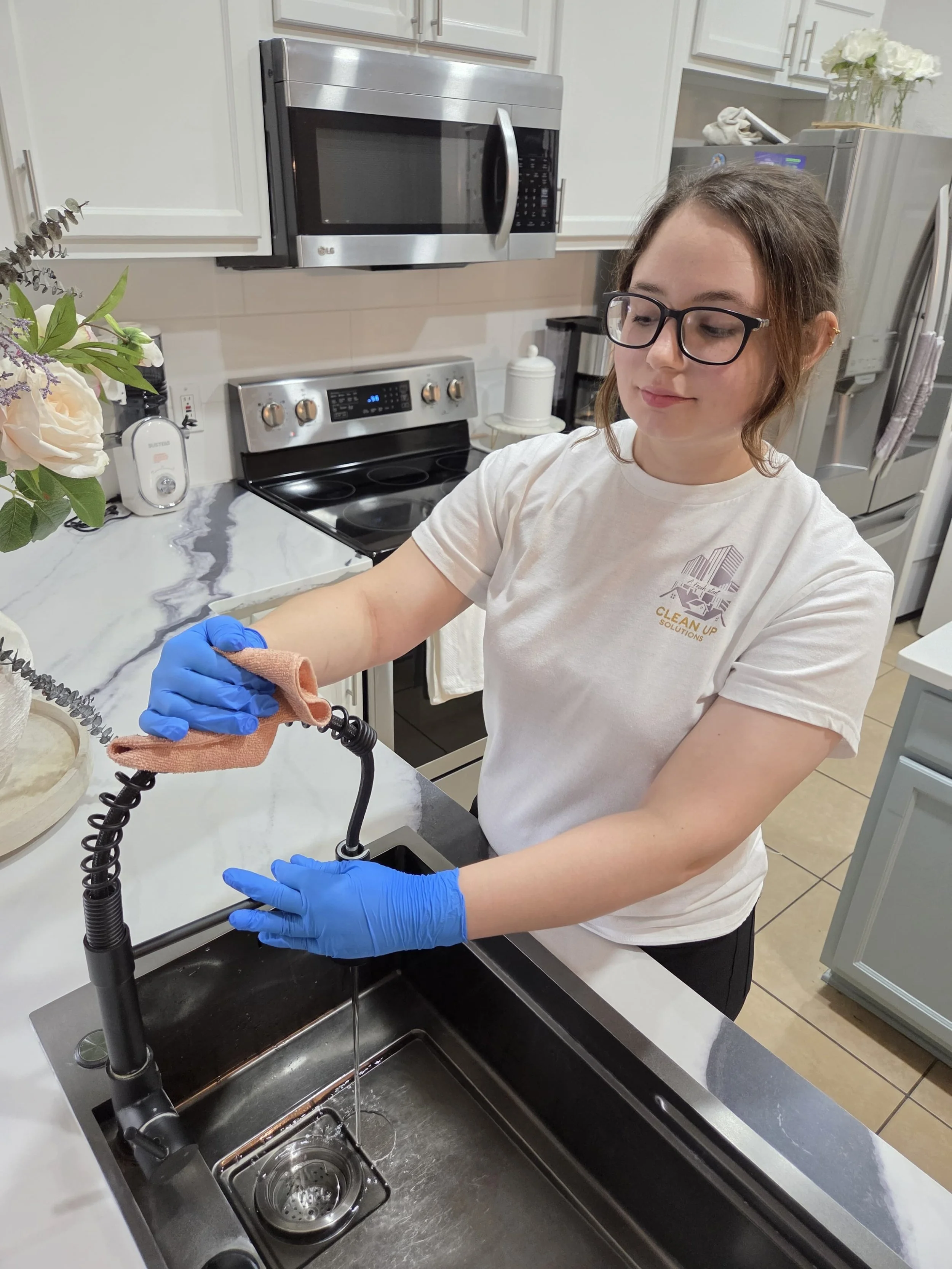 A woman wearing glasses, a white t-shirt with a logo, and blue gloves is disinfecting a kitchen sink with a cloth.