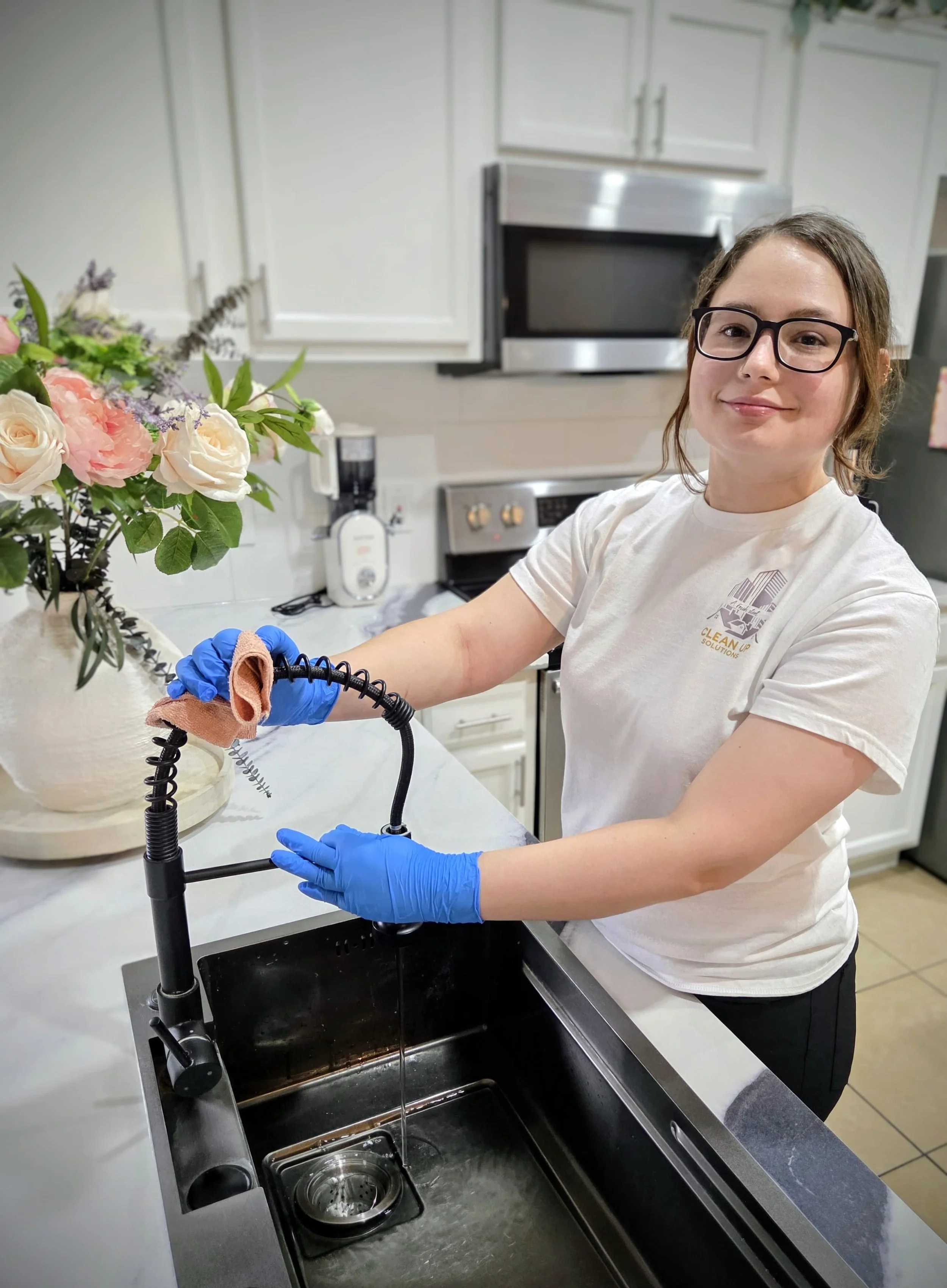 A woman with glasses and wearing blue gloves cleaning a kitchen sink with a spray nozzle and a cloth, standing next to a bouquet of flowers in a white vase on the counter.