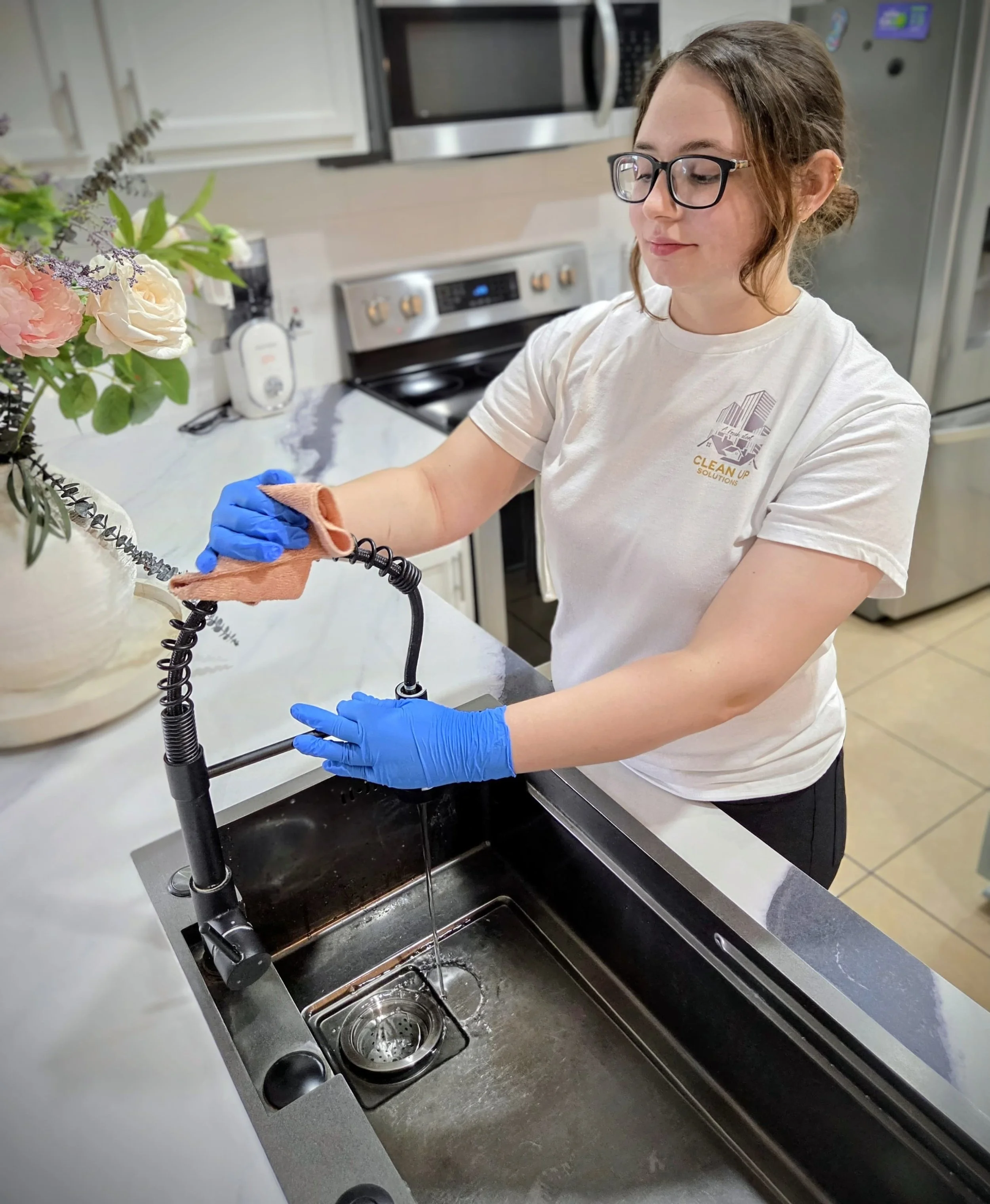 A woman wearing glasses, a white t-shirt, and blue gloves is cleaning a kitchen sink with a cloth. There is a vase of flowers nearby, and the kitchen has modern appliances like a microwave, oven, and refrigerator.